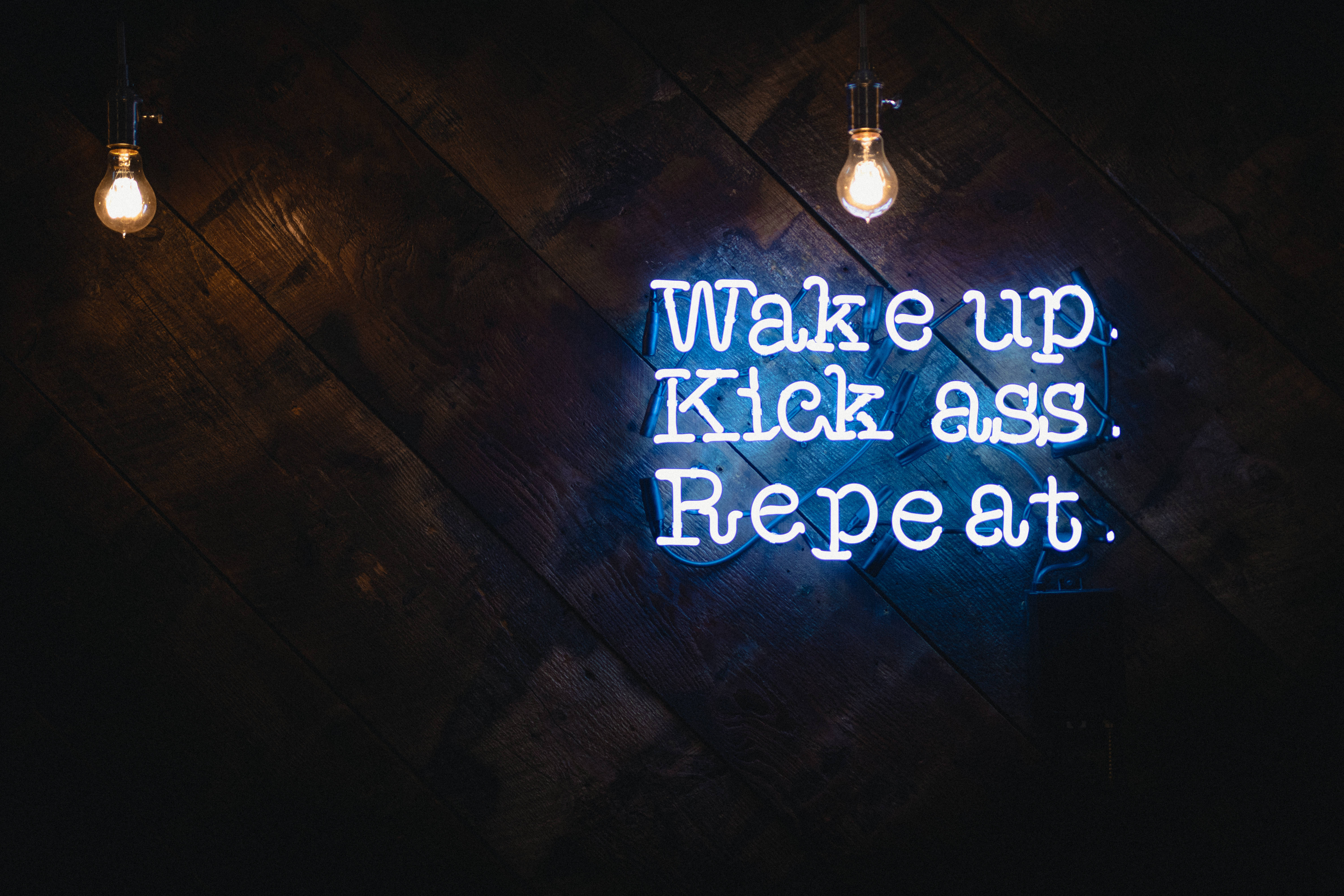 激勵 桌布 壁紙 — 一個發光的藍色霓虹燈牌，上面寫著「Wake up. Kick ass. Repeat.」，掛在深色木牆上，下方有兩個溫暖的吊燈。