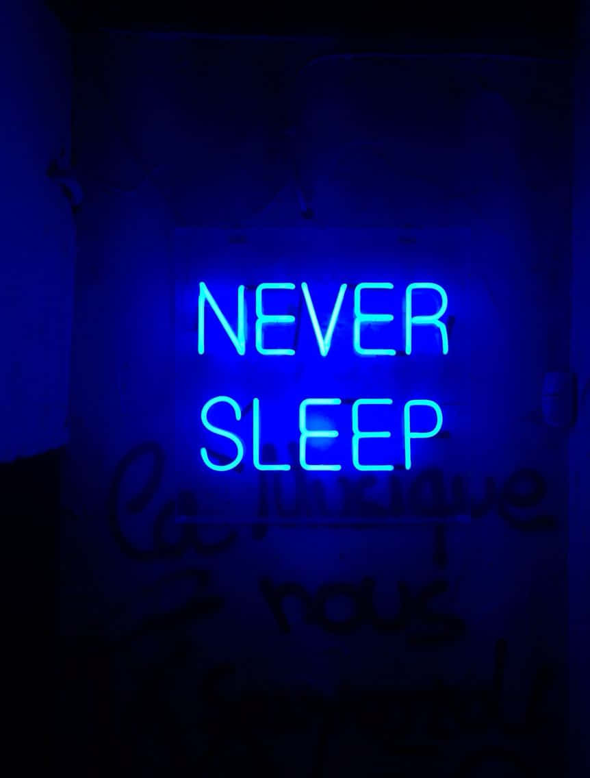 霓虹藍美學 桌布 壁紙 — 一個發光的藍色霓虹燈牌，上面寫著 NEVER SLEEP，照亮了一面帶有微弱塗鴉的暗牆。