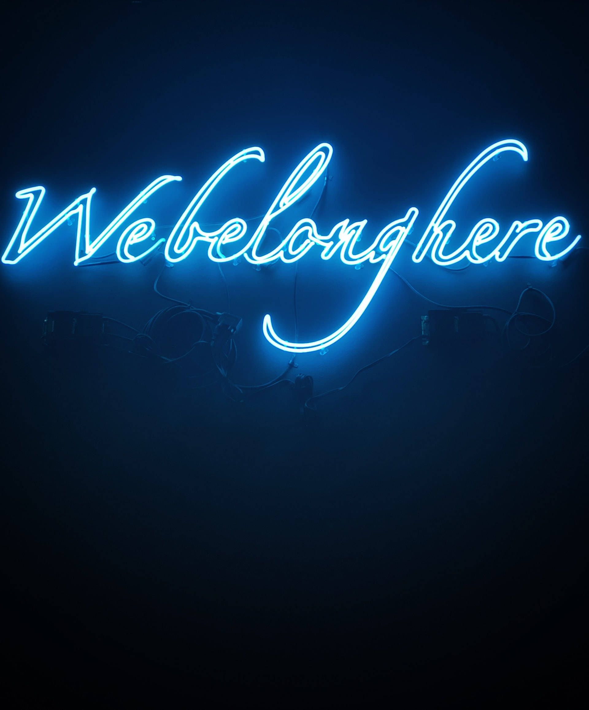 霓虹燈 桌布 壁紙 — 一個發光的藍色霓虹燈牌，上面寫著 'We belong here'，背景是深色牆壁，下方有可見的電線。