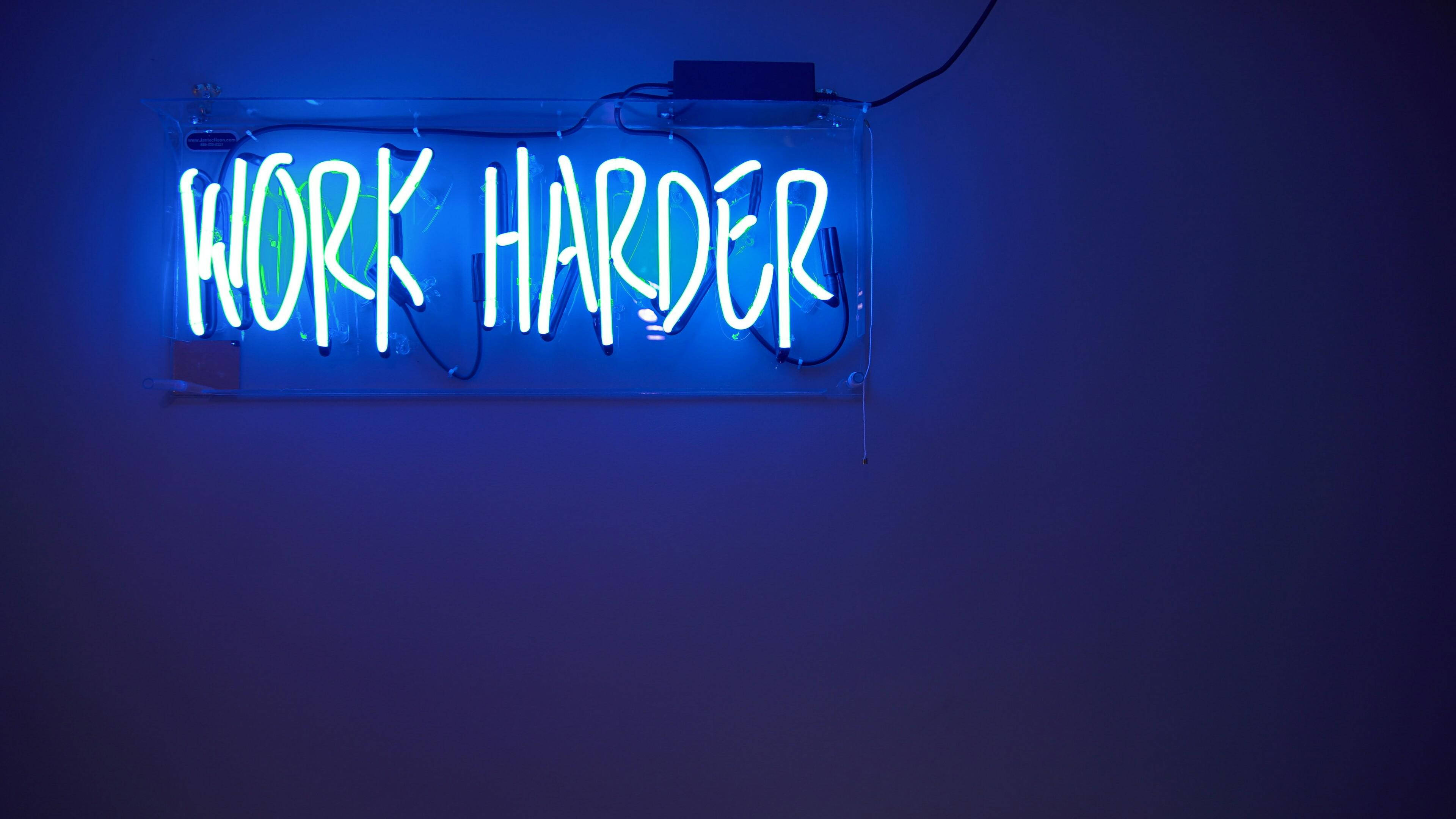 霓虹 桌布 壁紙 — 一個發光的藍色霓虹燈牌，上面寫著「WORK HARDER」，安裝在深色牆壁上，投射出充滿活力的電光色調。