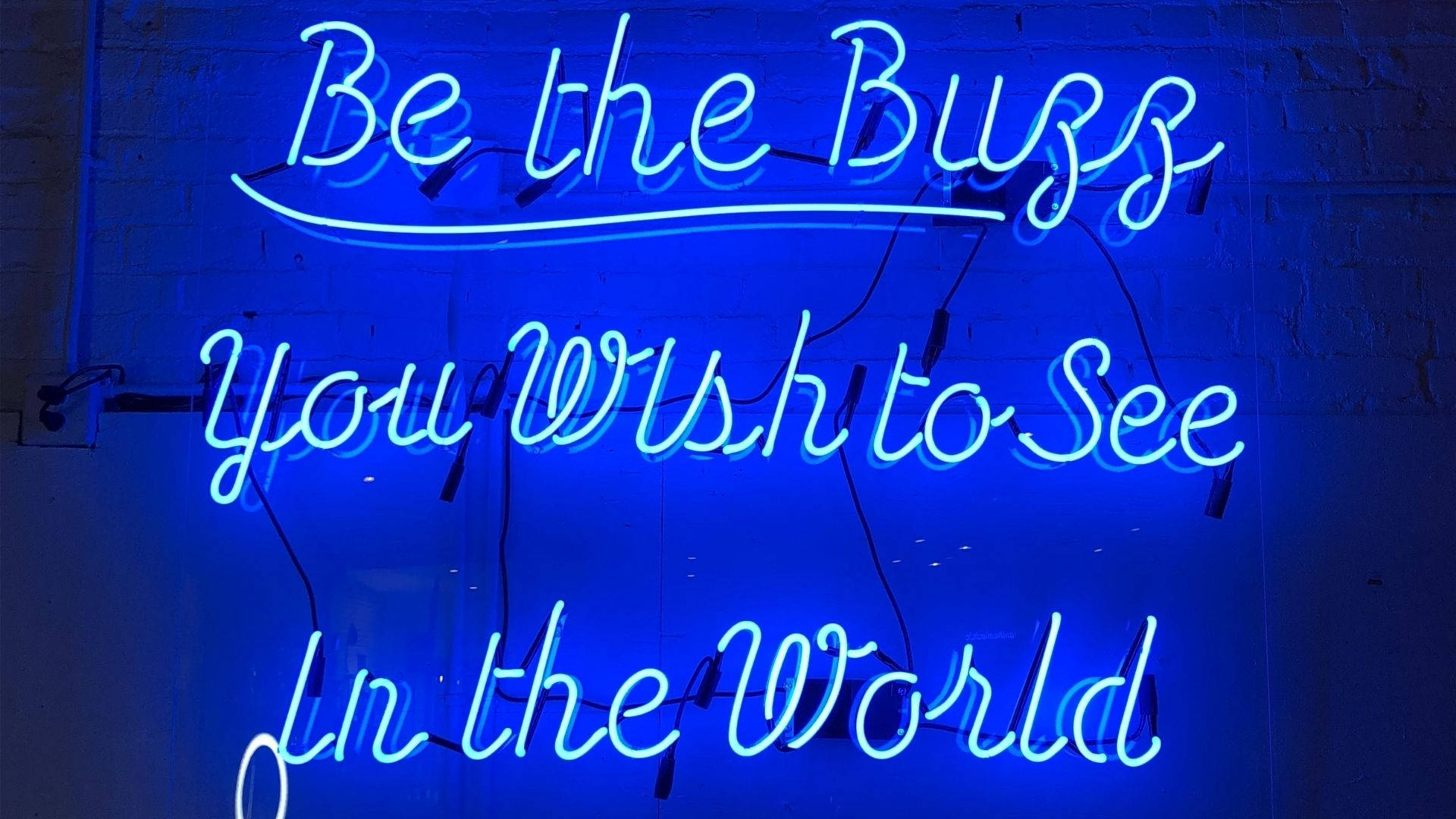 霓虹藍動畫 桌布 壁紙 — 一個發光的藍色霓虹燈牌，上面寫著 'Be the Buzz You Wish to See In the World'，背景是深色磚牆。