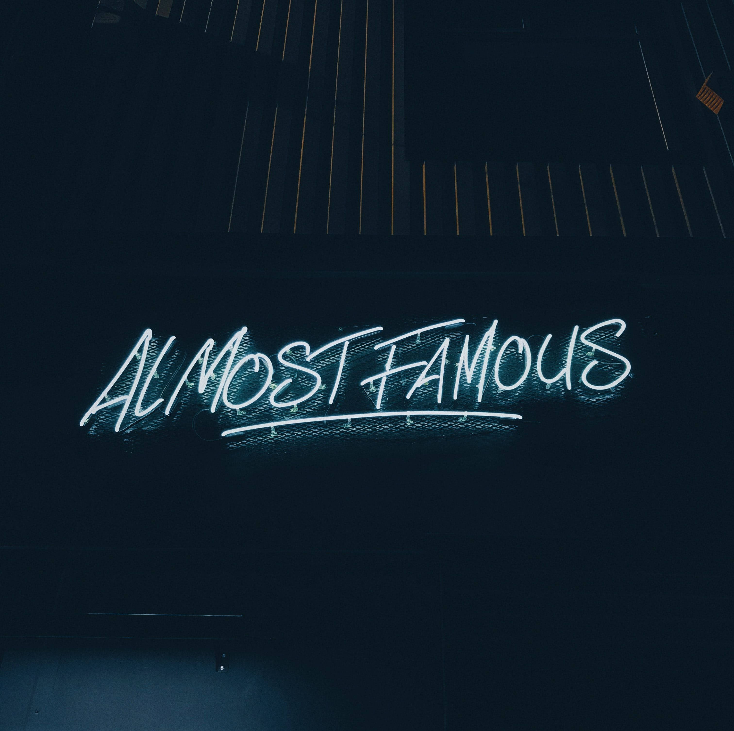 Digital 桌布 壁紙 — 一個發光的藍色霓虹燈牌，上面寫著「ALMOST FAMOUS」，在夜晚照亮了黑暗的城市牆壁。