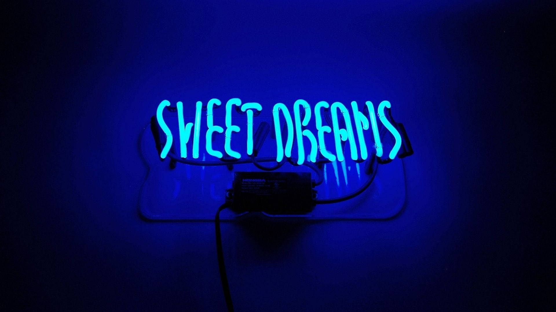霓虹藍動畫 桌布 壁紙 — 一個發光的藍色霓虹燈牌，上面寫著 sweet dreams，在昏暗的房間中將柔和的光線投射在深色牆壁上。