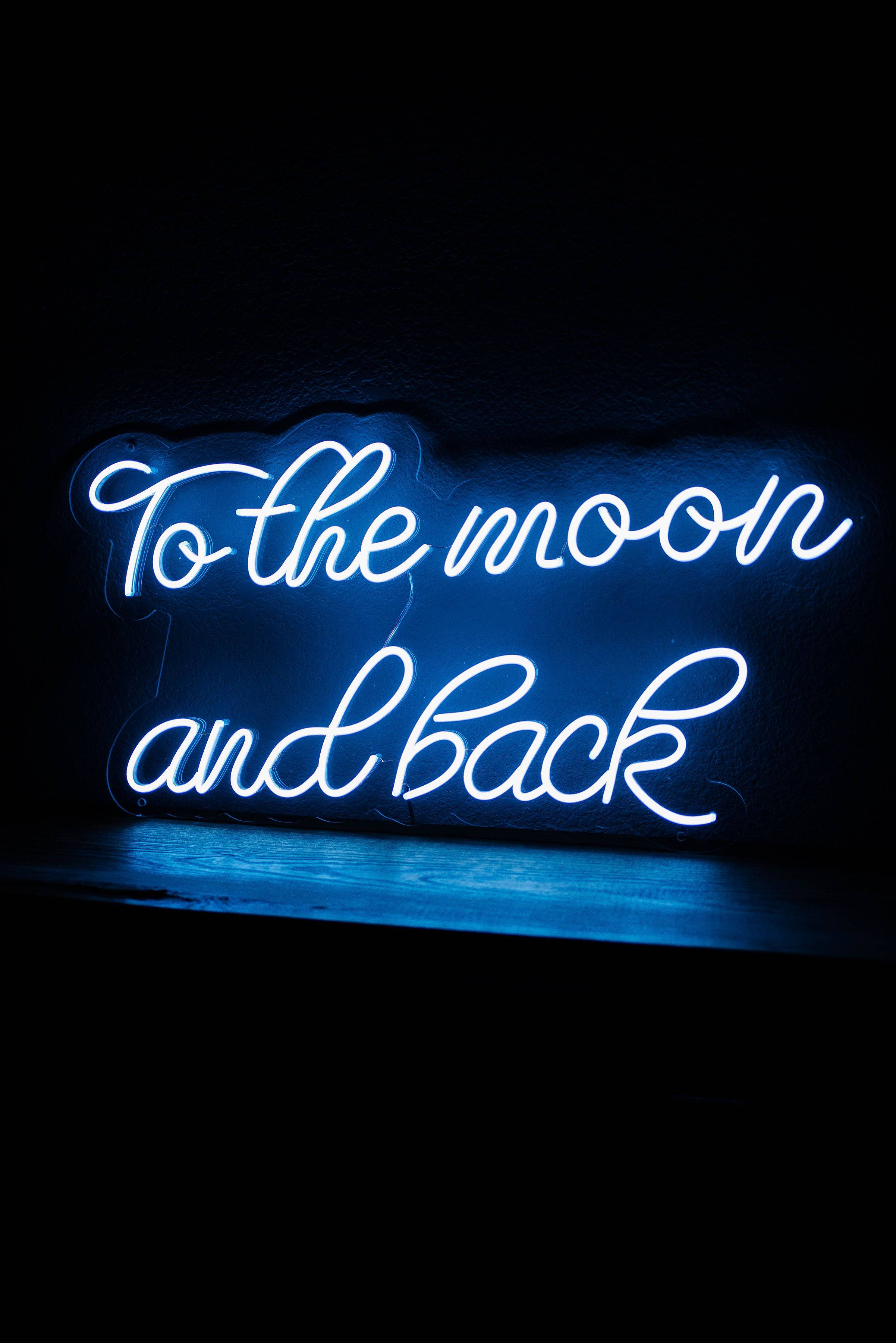 霓虹藍美學 桌布 壁紙 — 一個發光的藍色霓虹燈牌，上面寫著 'To the moon and back'，背景是深色紋理牆壁，下方有反射表面。