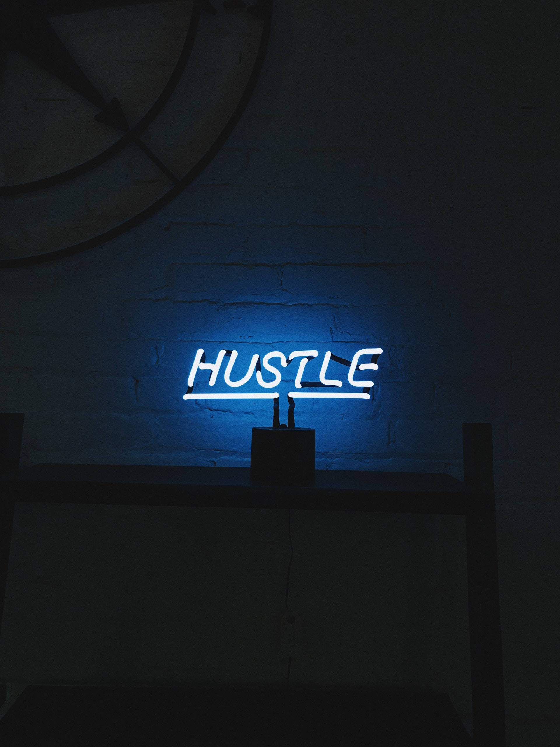 Hustle 桌布 壁紙 — 一個發光的藍色霓虹燈牌，拼寫著 HUSTLE，照亮了陰暗磚牆，營造出憂鬱、極簡的室內氛圍。