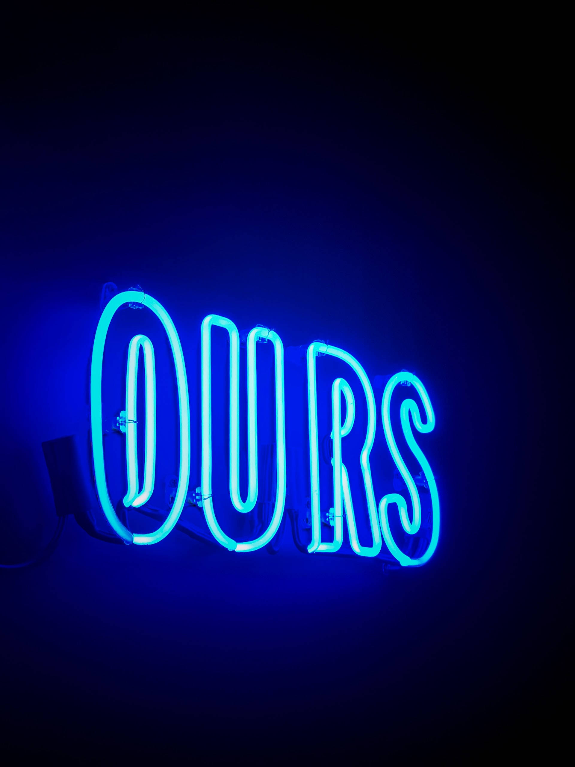 霓虹燈 桌布 壁紙 — 一個發光的藍色霓虹燈牌，拼寫著「OURS」，在極簡環境中對著深色牆壁投射出鮮豔的光芒。