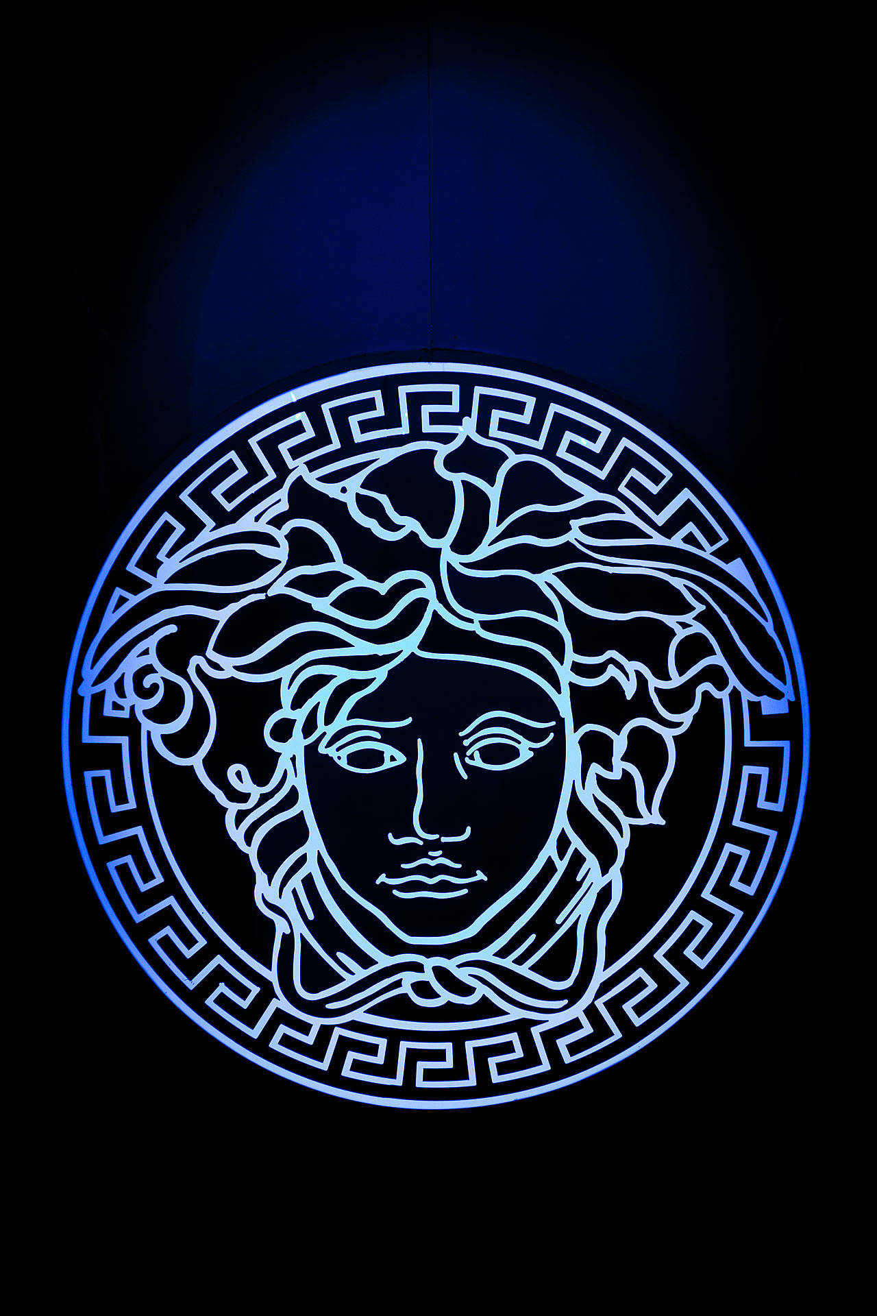 Versace Logo 桌布 壁紙 — 一個發光的藍色霓虹燈牌，描繪了美杜莎頭像，帶有蛇髮和希臘鑰匙邊框，背景為深色。