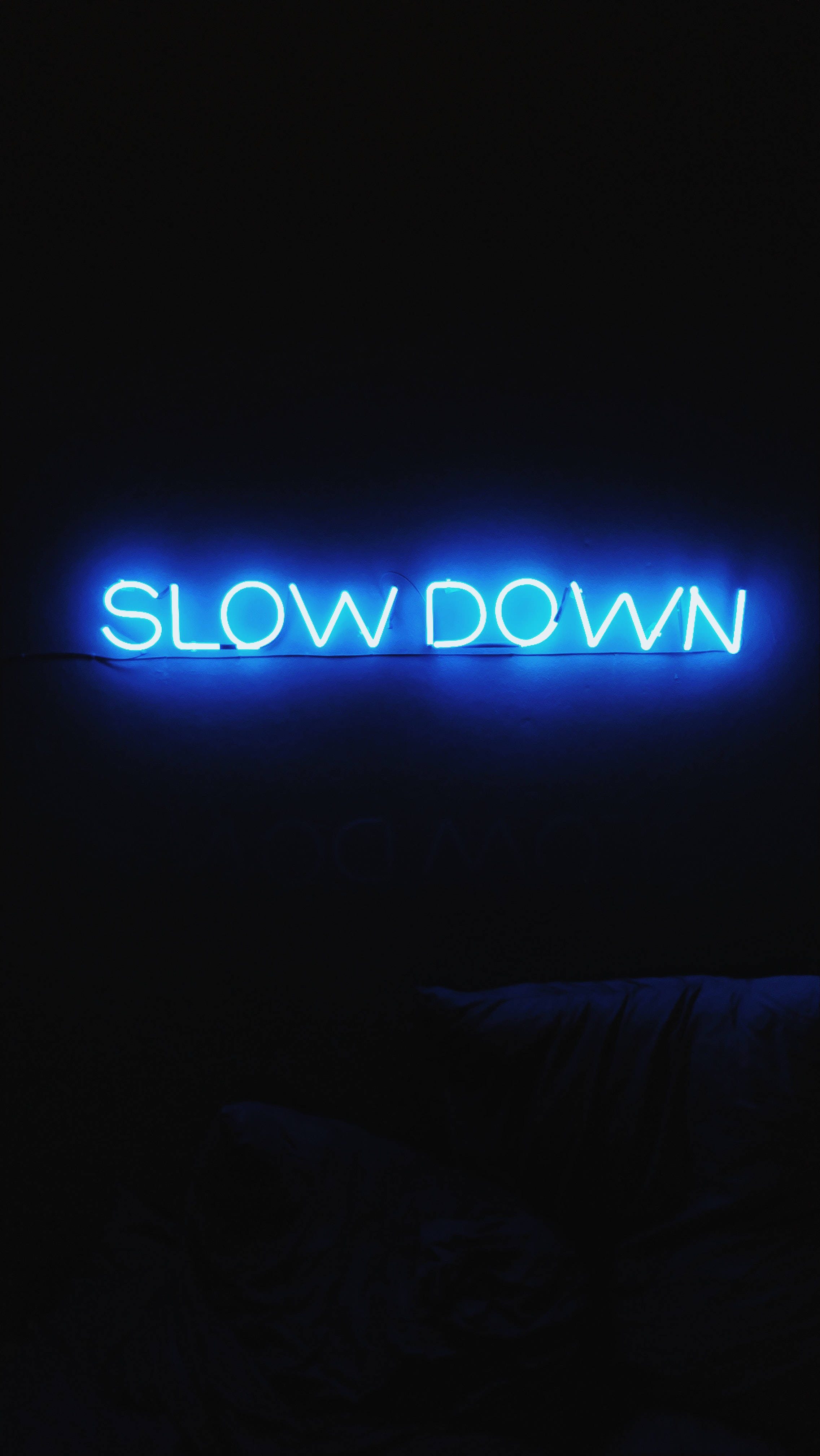 neon sign 桌布 壁紙 — 一個發光的藍色霓虹燈牌寫著 slow down，照亮了下方有床的黑暗房間。