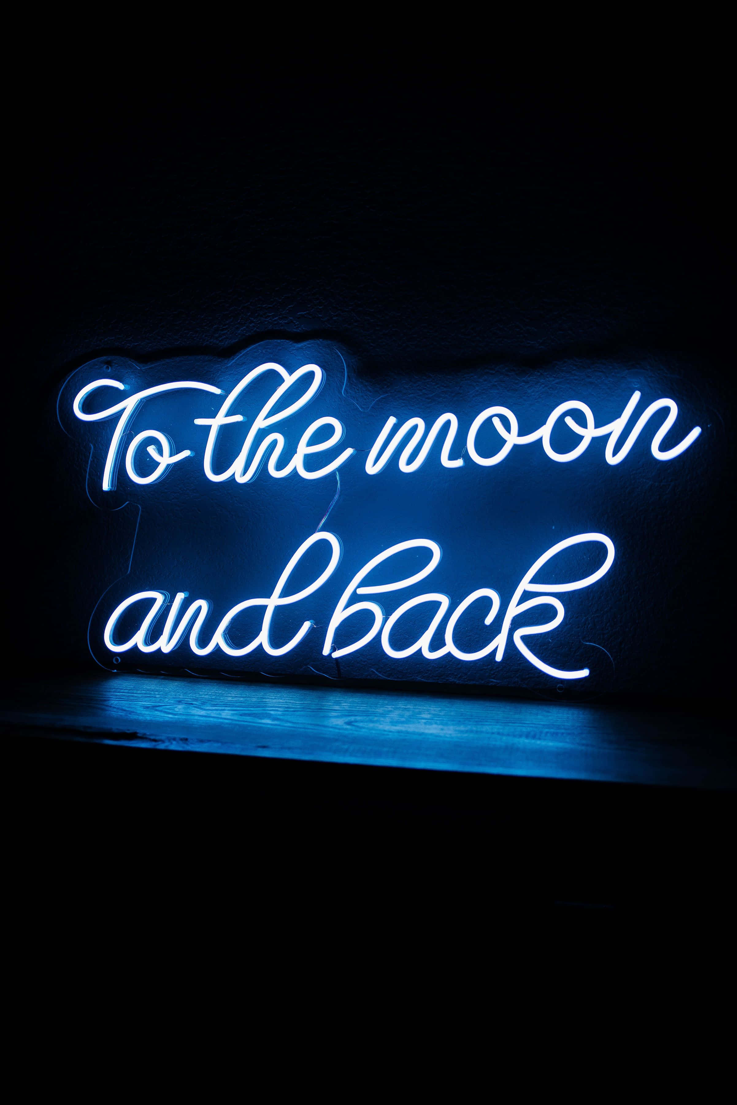 霓虹藍美學 桌布 壁紙 — 一個發光的藍色霓虹燈牌寫著「To the moon and back」，背景是深色紋理牆壁，下方有反光表面。