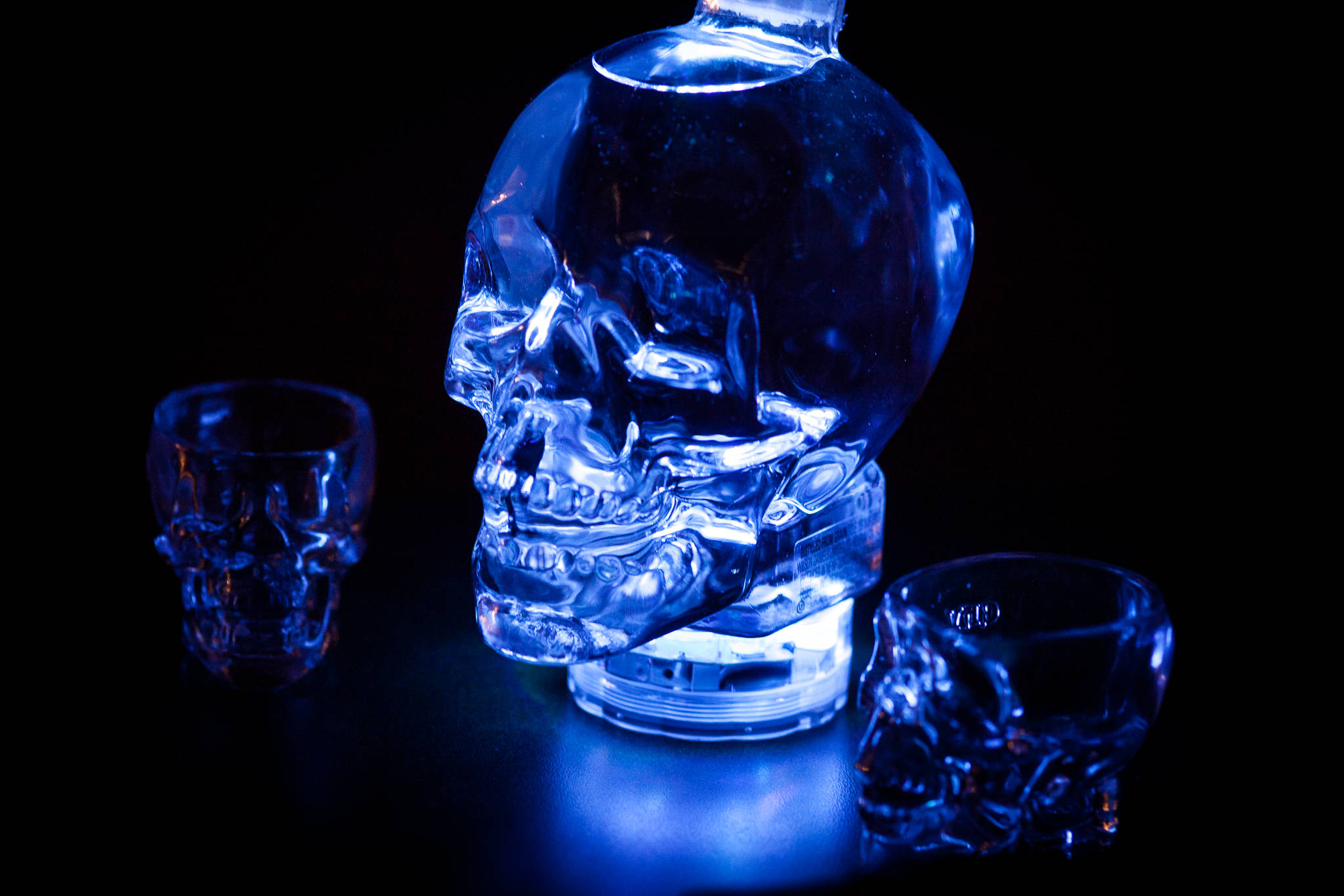 Drink 桌布 壁紙 — 一個發光的藍色骷髏形狀瓶子和兩個相配的骷髏杯，在黑色背景下照亮。