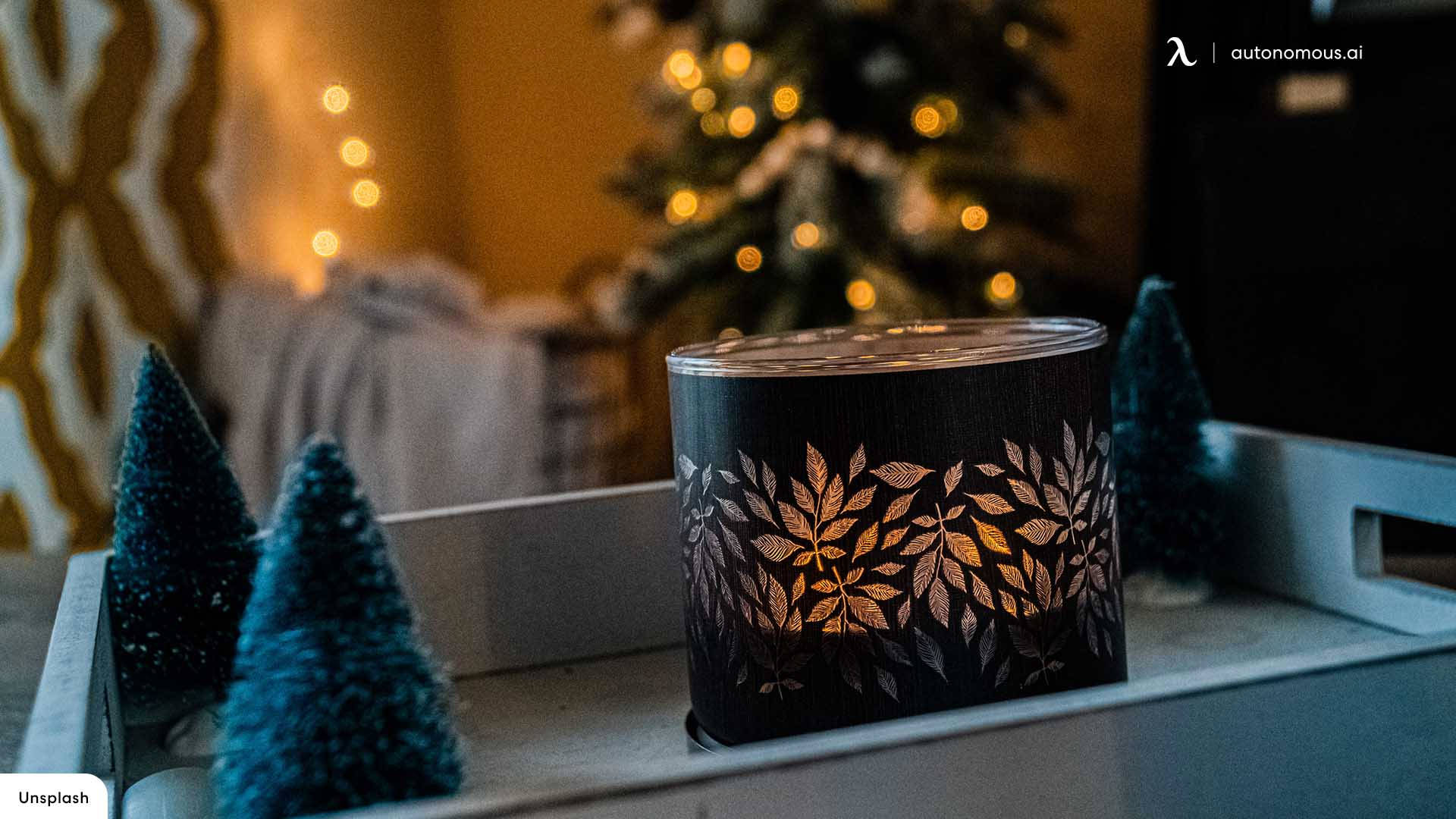 Cozy 桌布 壁紙 — 一個發光的蠟燭放在黑色葉紋燭台上，置於托盤上，旁邊有迷你藍色樹木，背景是模糊的聖誕樹和溫暖的燈光。
