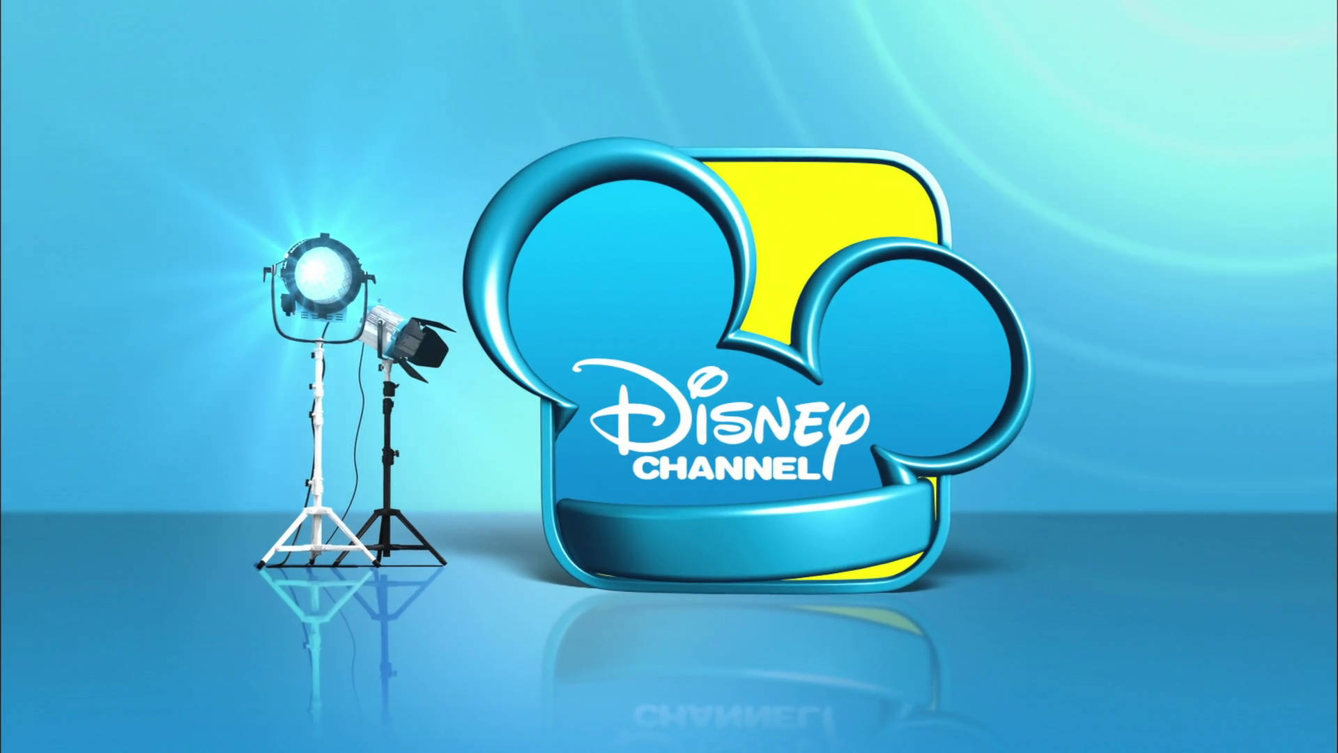 Disney Channel 桌布 壁紙 — 一個發光的迪士尼頻道標誌，帶有米奇老鼠耳朵，旁邊有一個攝影棚聚光燈，位於反光的藍色表面上。