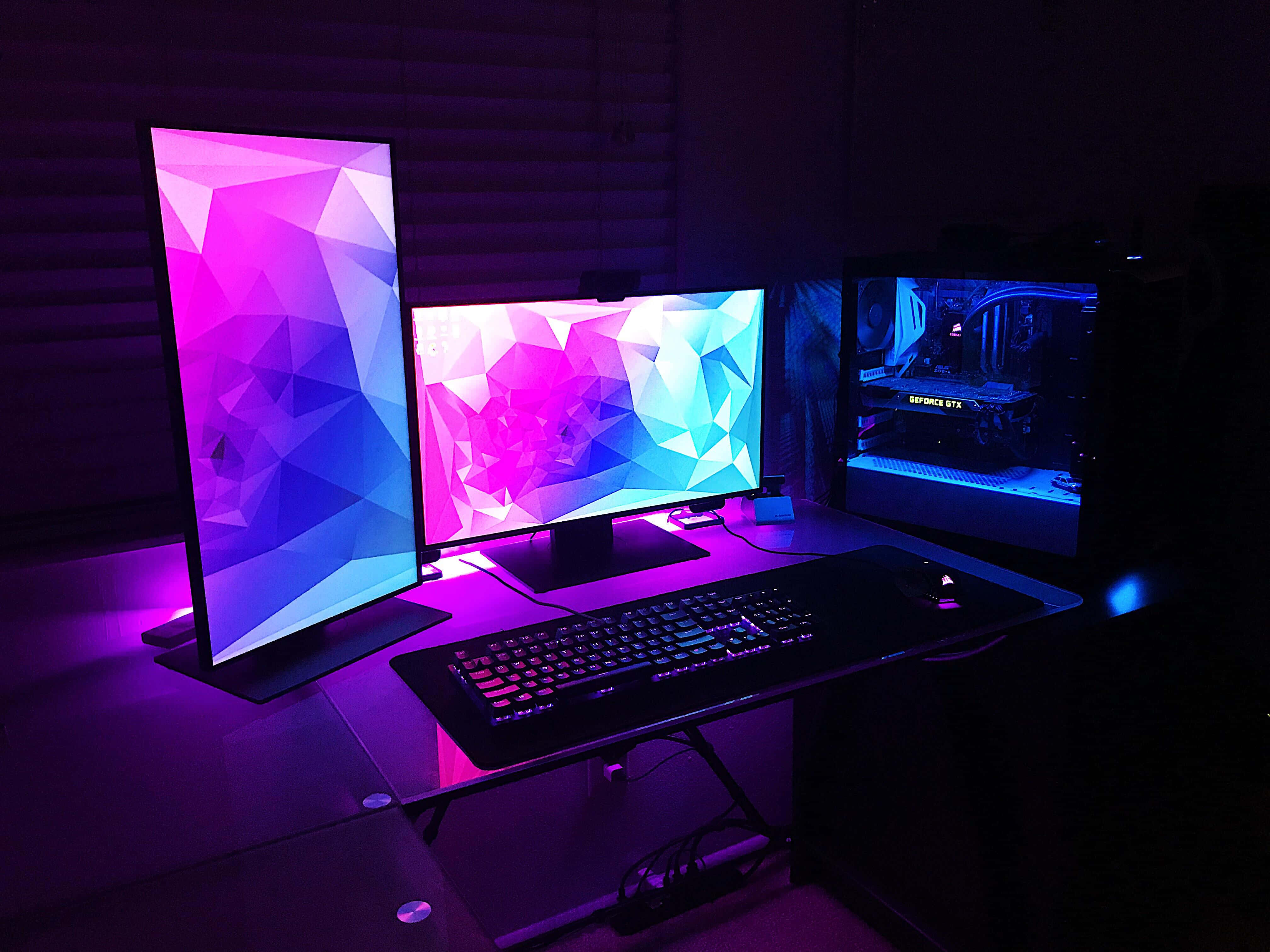 Gaming Setup 桌布 壁紙 — 一個發光的遊戲設置，配有三個顯示器，顯示鮮豔的幾何桌布，以及在黑暗房間中發光的電腦機箱。