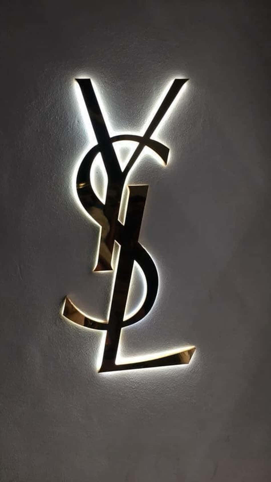 Ysl 桌布 壁紙 — 一個發光的金色YSL字母組合標誌，帶有背光，安裝在紋理灰色牆壁上。