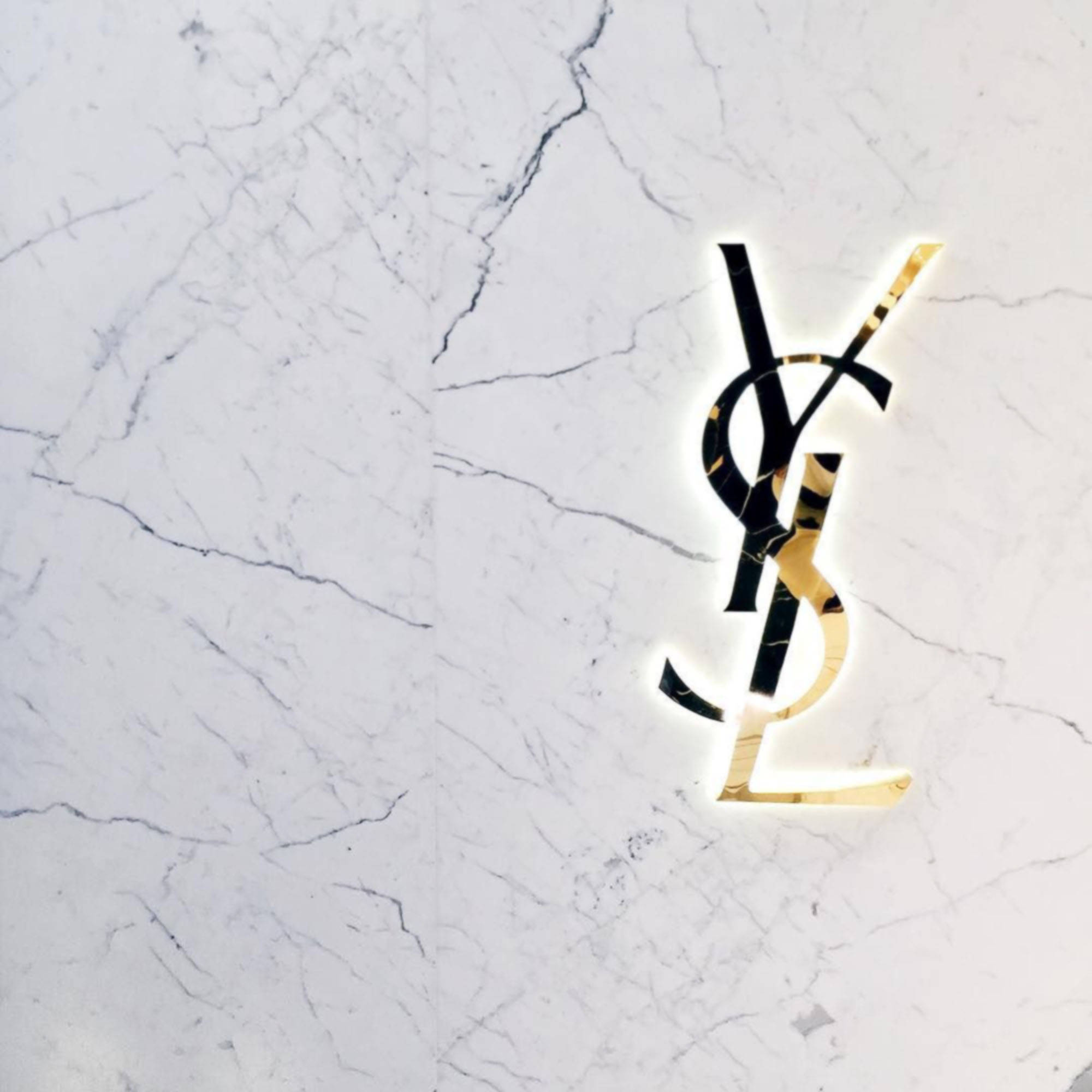 Ysl 桌布 壁紙 — 一個發光的金色和黑色YSL標誌嵌入帶有深灰色紋理的白色大理石中。