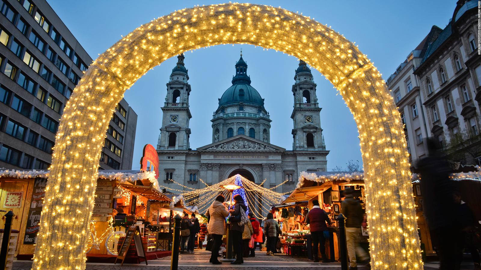 budapest 桌布 壁紙 — 一個發光的金色拱門框住了熱鬧的聖誕市集，人們在暮色中於宏偉大教堂前閃爍的燈光下購物。