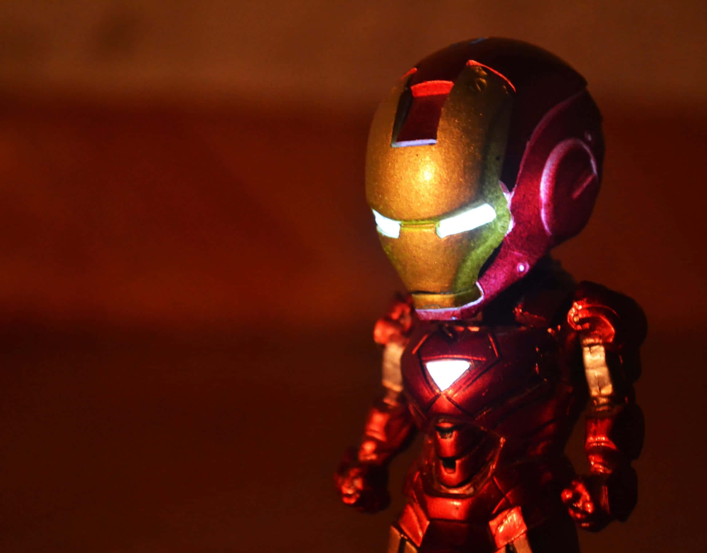 Iron Man Action Figures 桌布 壁紙 — 一個發光的鋼鐵人人偶站在昏暗燈光下，眼睛和胸部弧形反應器發光，背景溫暖模糊。
