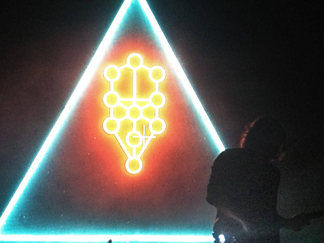 Frank Ocean 桌布 壁紙 — 一個發光的霓虹三角形帶有幾何符號照亮黑暗的舞台，一個剪影人物在觀看。