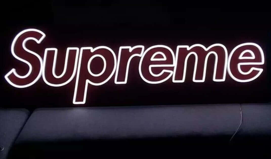 supreme 桌布 壁紙 — 一個發光的霓虹燈 Supreme 標誌，酒紅色和白色，在黑暗背景上投射出明亮的光線，下方有一張黑色沙發。