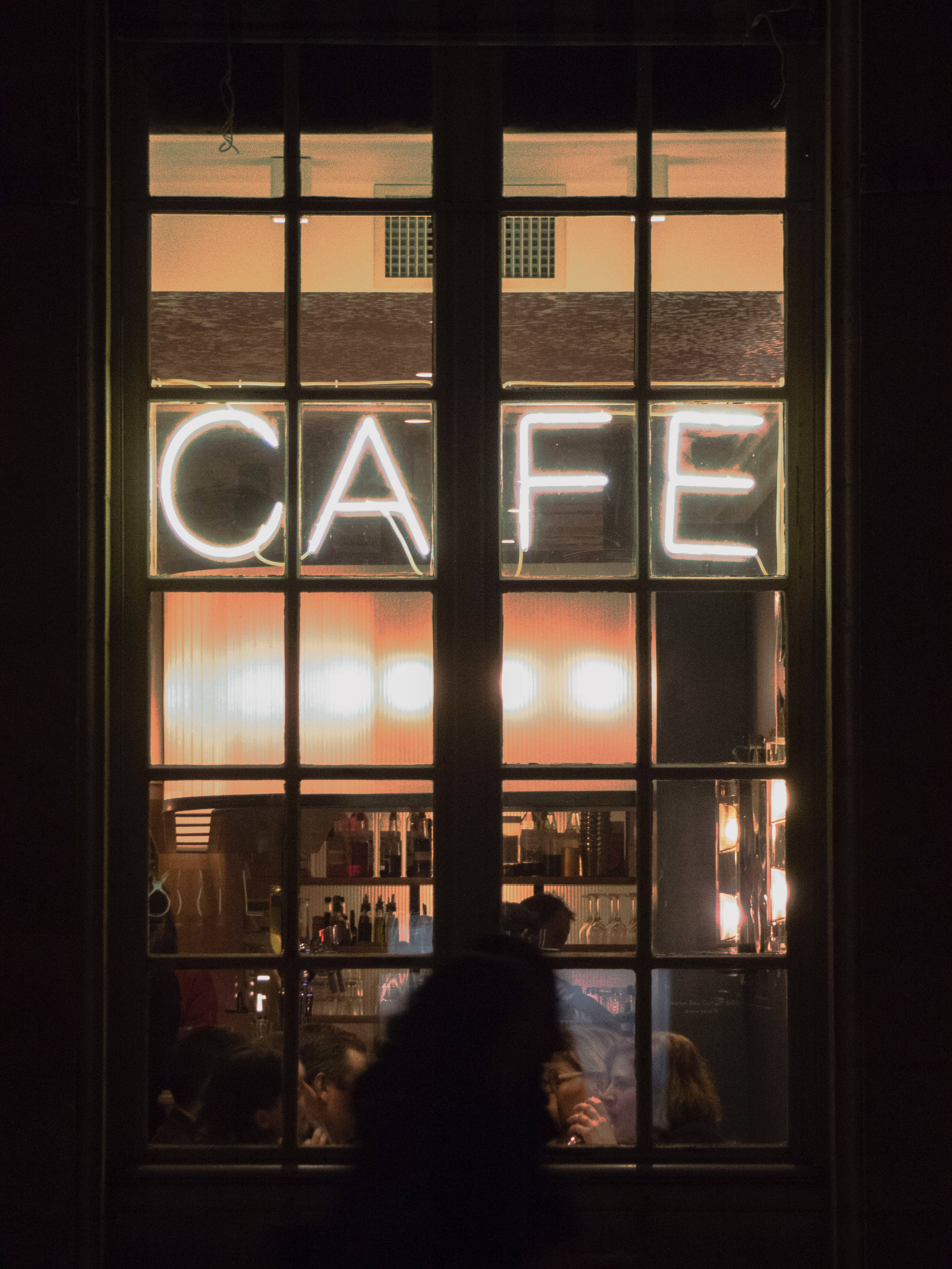 Coffee Shop 桌布 壁紙 — 一個發光的霓虹燈咖啡館招牌透過深色框窗戶照亮了舒適的室內，剪影般的顧客在裡面享受溫暖的氛圍。