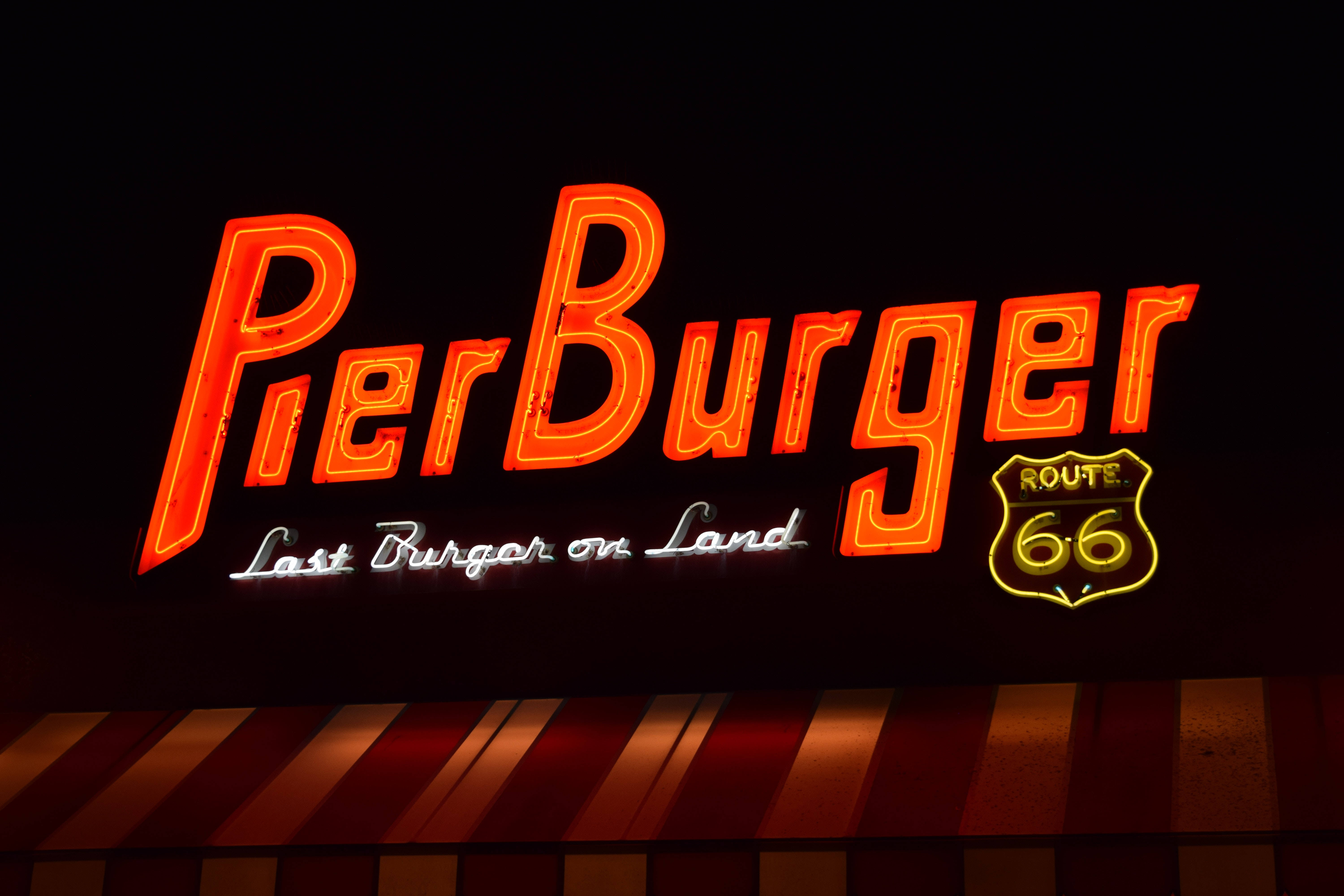 Monica 桌布 壁紙 — 一個發光的霓虹燈招牌，上面有Pier Burger和Route 66標誌，在黑暗的夜空下，位於條紋遮陽篷上方閃耀。