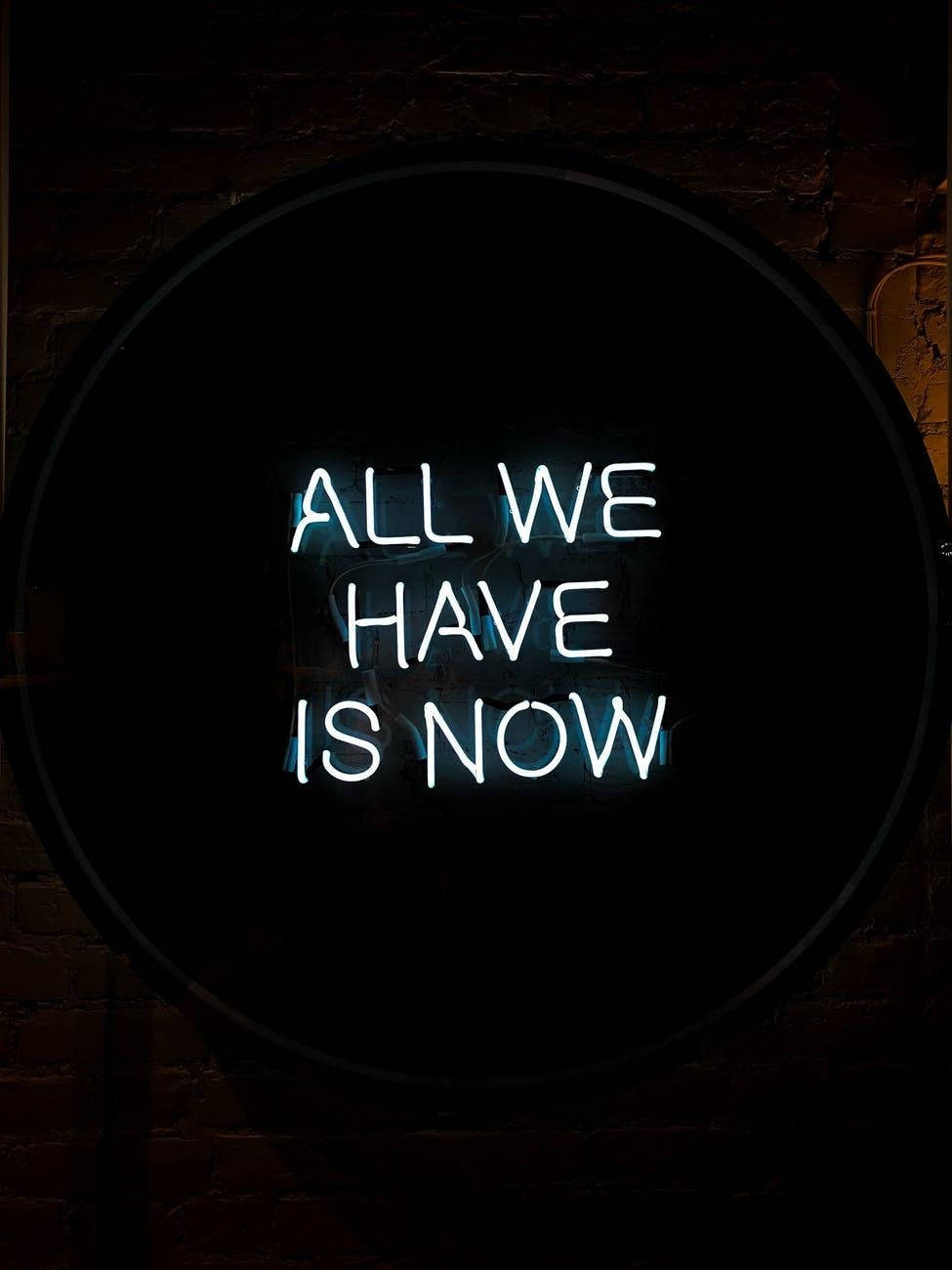 Motivational Quotes 桌布 壁紙 — 一個發光的霓虹燈牌，上面寫著「ALL WE HAVE IS NOW」，放在圓形框架內，背景是深色磚牆。