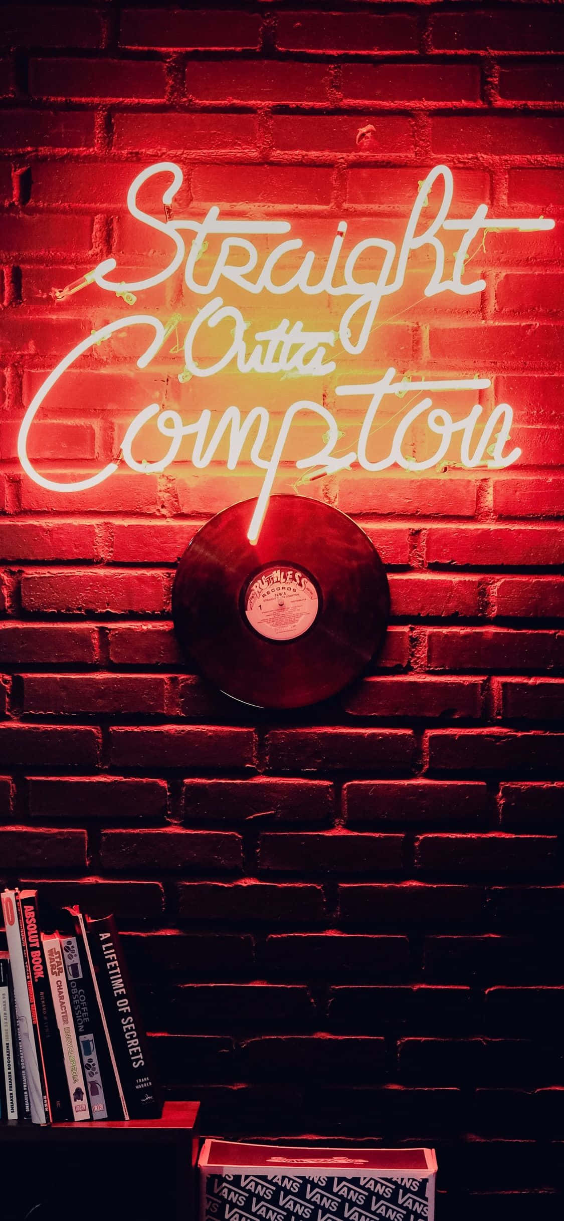 iPhone Neon 桌布 壁紙 — 一個發光的霓虹燈牌，上面寫著「Straight Outta Compton」，在磚牆上投射出紅光，旁邊有一張黑膠唱片和書籍。