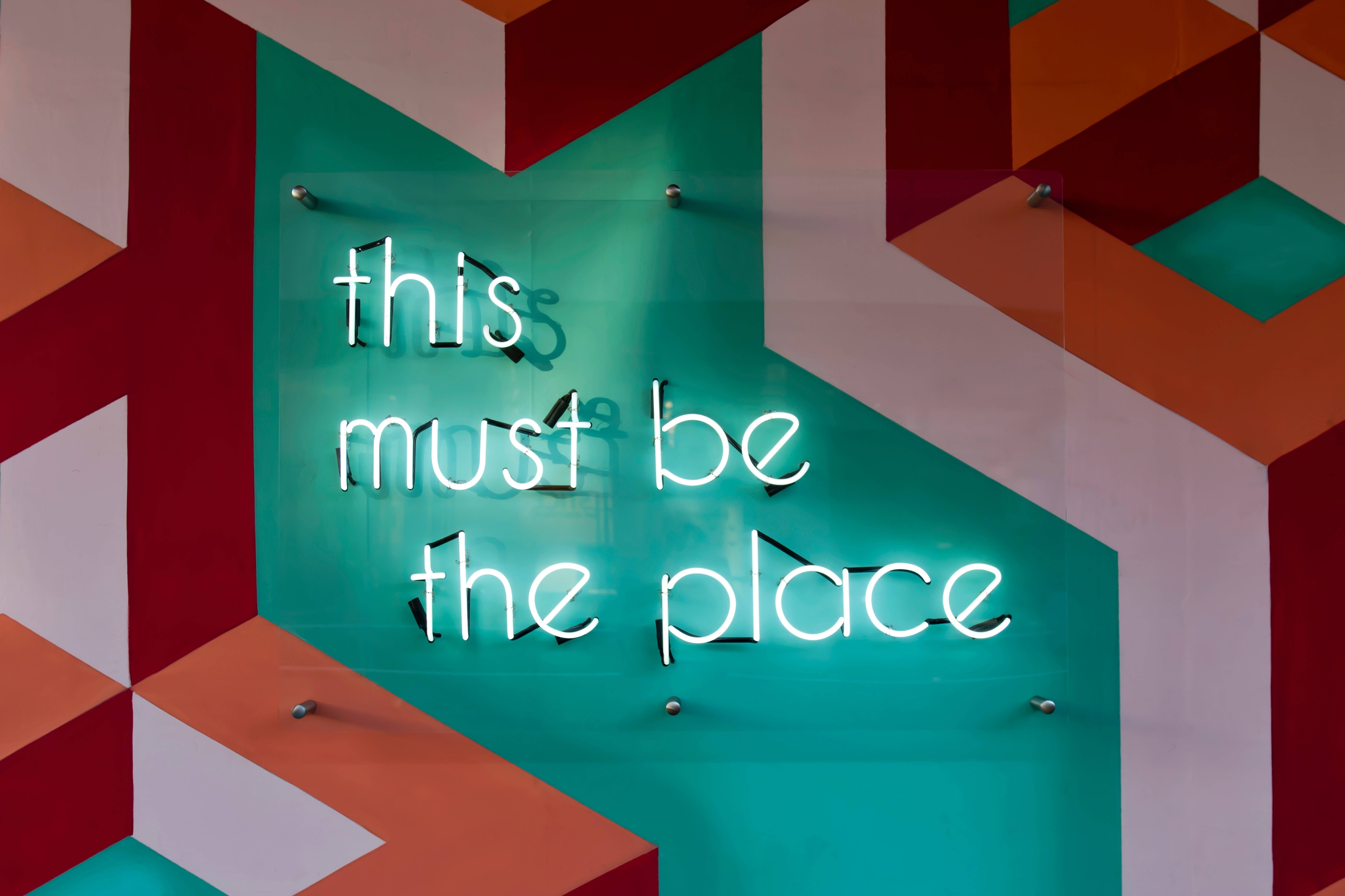 幾何 桌布 壁紙 — 一個發光的霓虹燈牌，上面寫著「this must be the place」，安裝在色彩繽紛的幾何牆上。