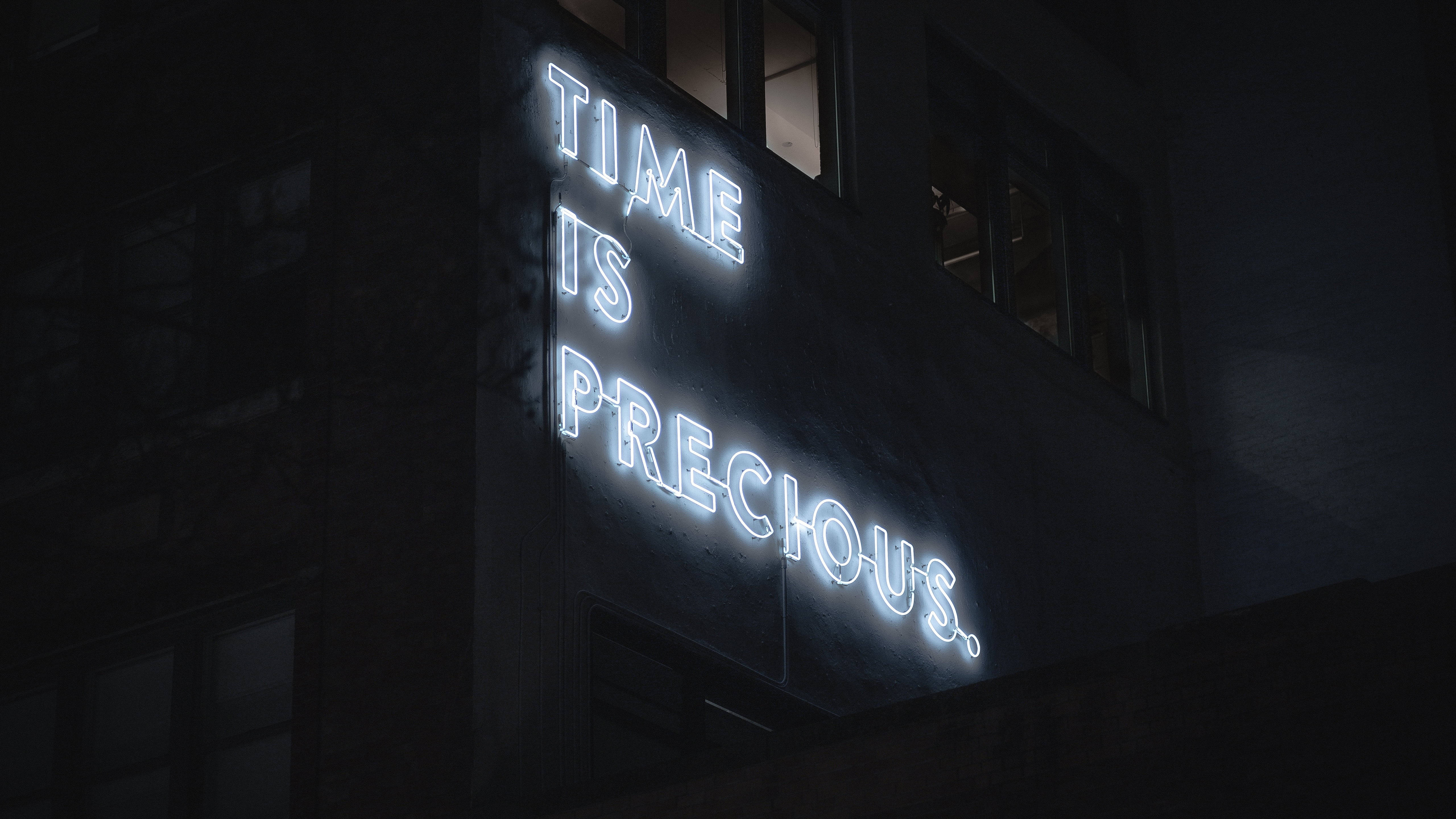 霓虹 桌布 壁紙 — 一個發光的霓虹燈牌，上面寫著 time is precious，在夜晚照亮了黑暗的城市建築外牆。