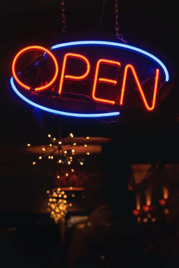 桌布 壁紙 — 一個發光的霓虹燈牌，用紅色字母拼出OPEN，帶有藍色橢圓形邊框，懸掛在黑暗中，下方是柔和照明的室內。