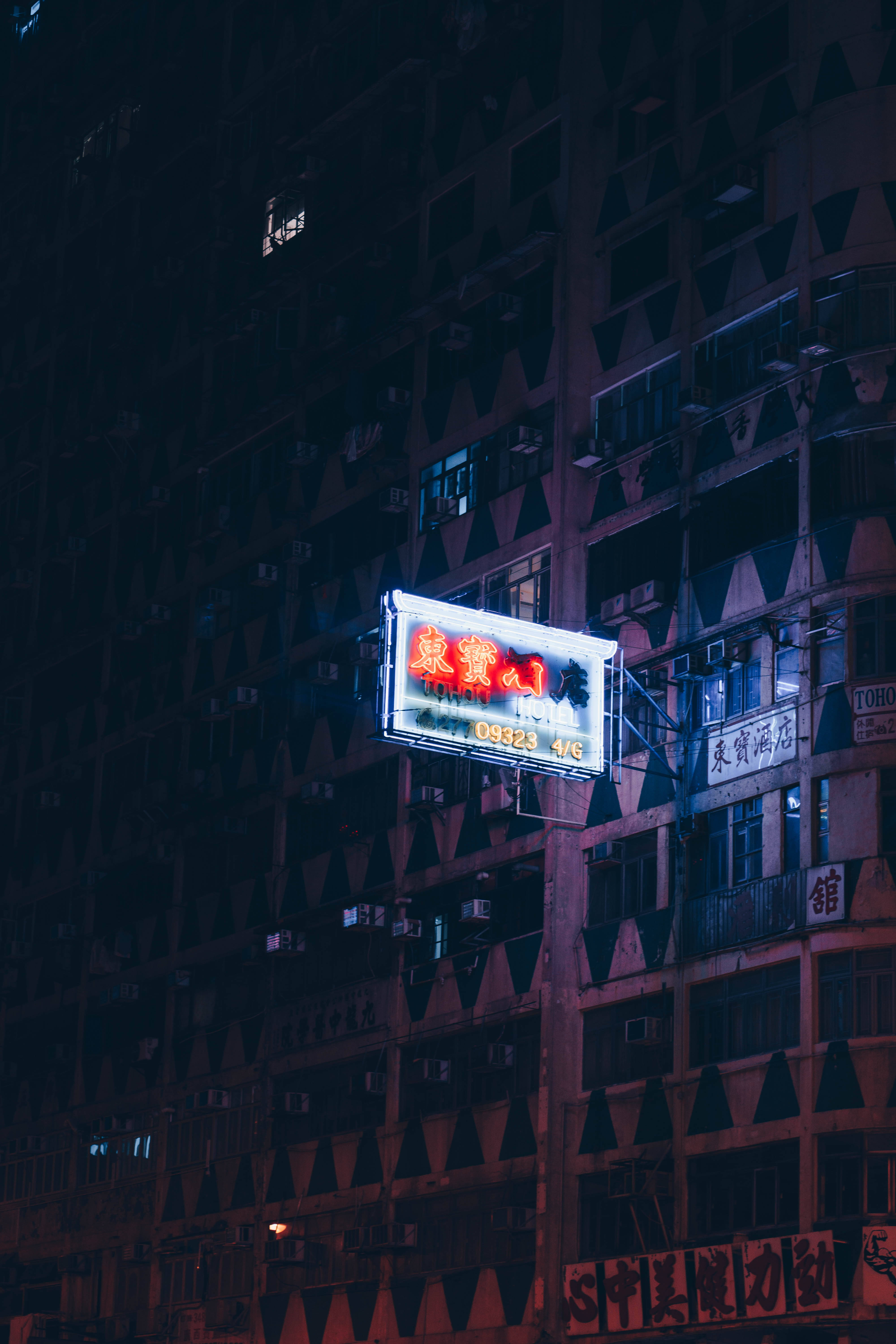 Neon Phone 桌布 壁紙 — 一個發光的霓虹燈牌在夜晚照亮了密集的城市建築外牆，將溫暖的光線投射在黑暗的幾何圖案上。