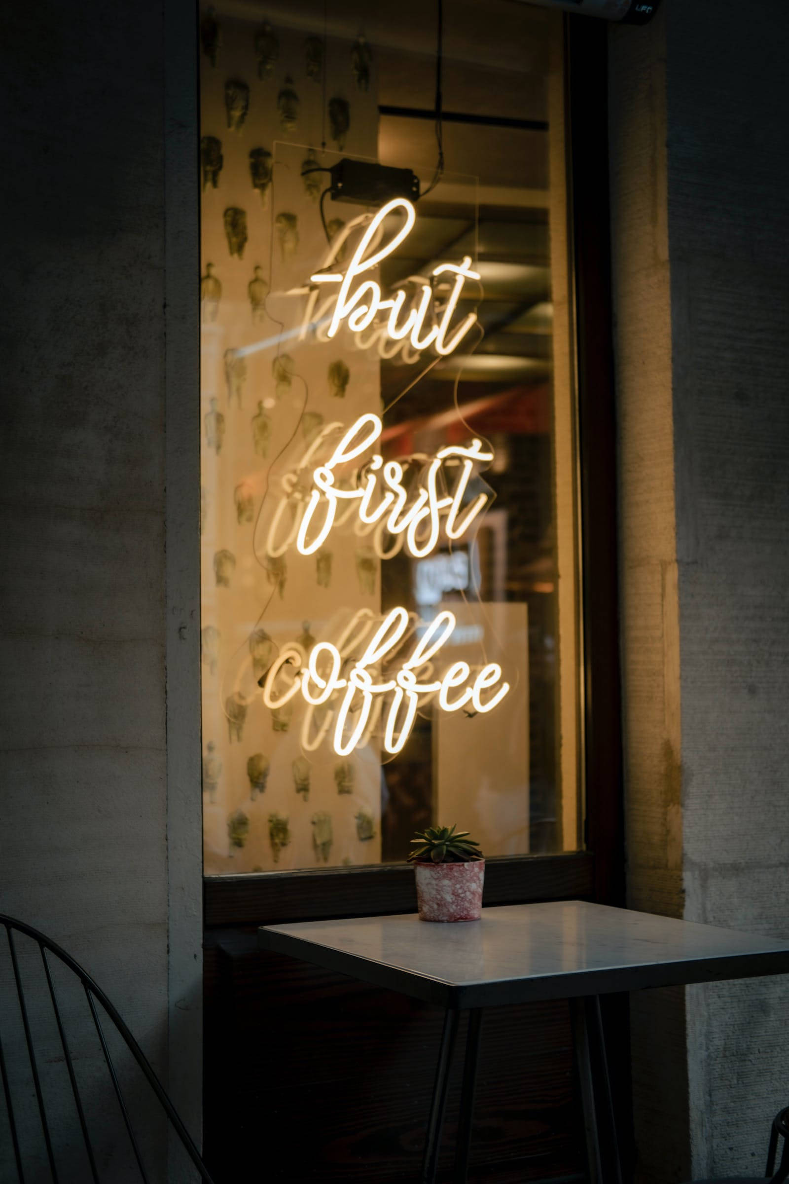 Neon Phone 桌布 壁紙 — 一個發光的霓虹燈牌寫著「but first coffee」，照亮了夜間舒適的咖啡館窗戶，外面桌子上放著一盆小盆栽。