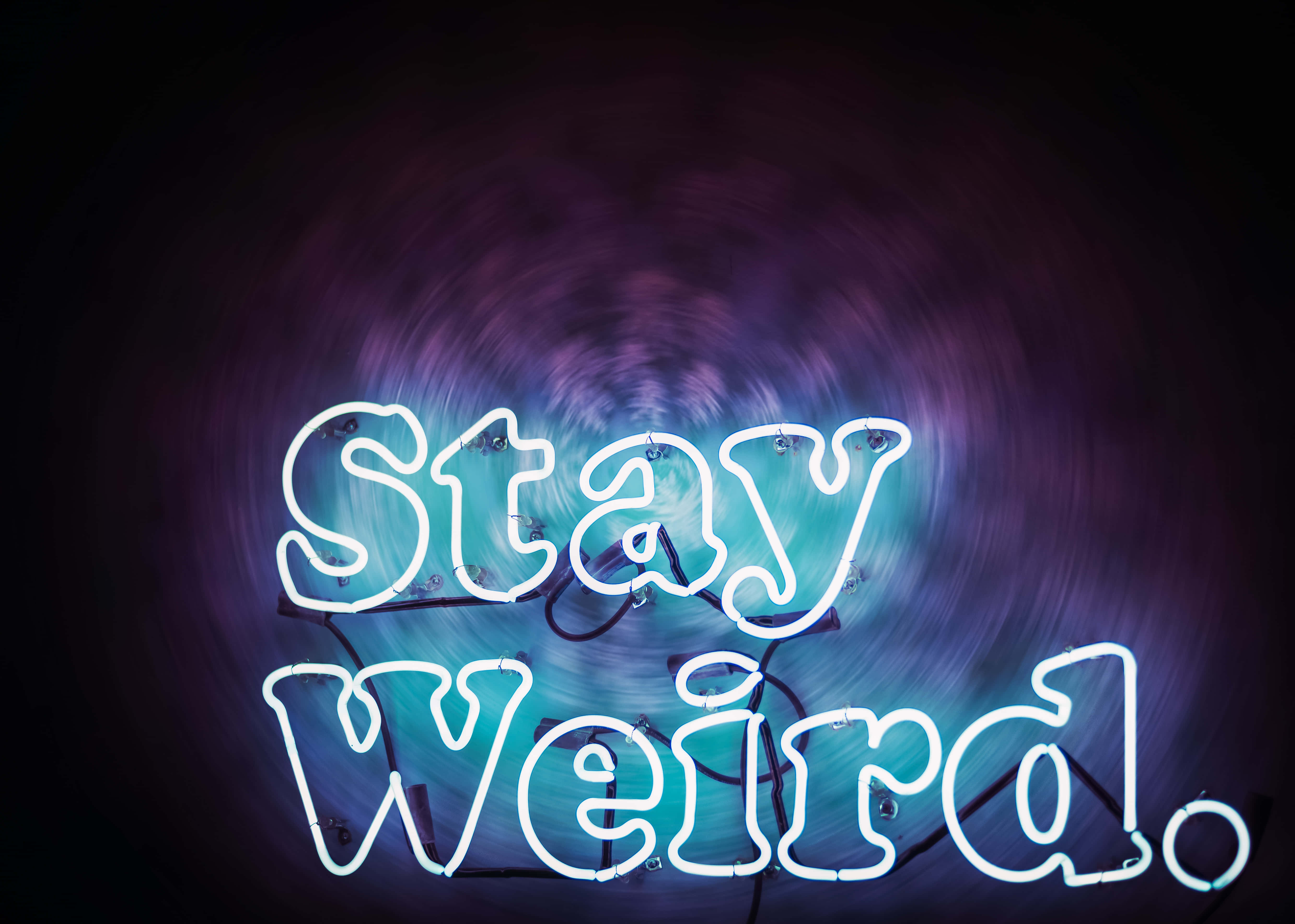 美學語錄動圖 桌布 壁紙 — 一個發光的霓虹燈牌寫著「Stay Weird.」，背景是旋轉的紫色和藍色漩渦。