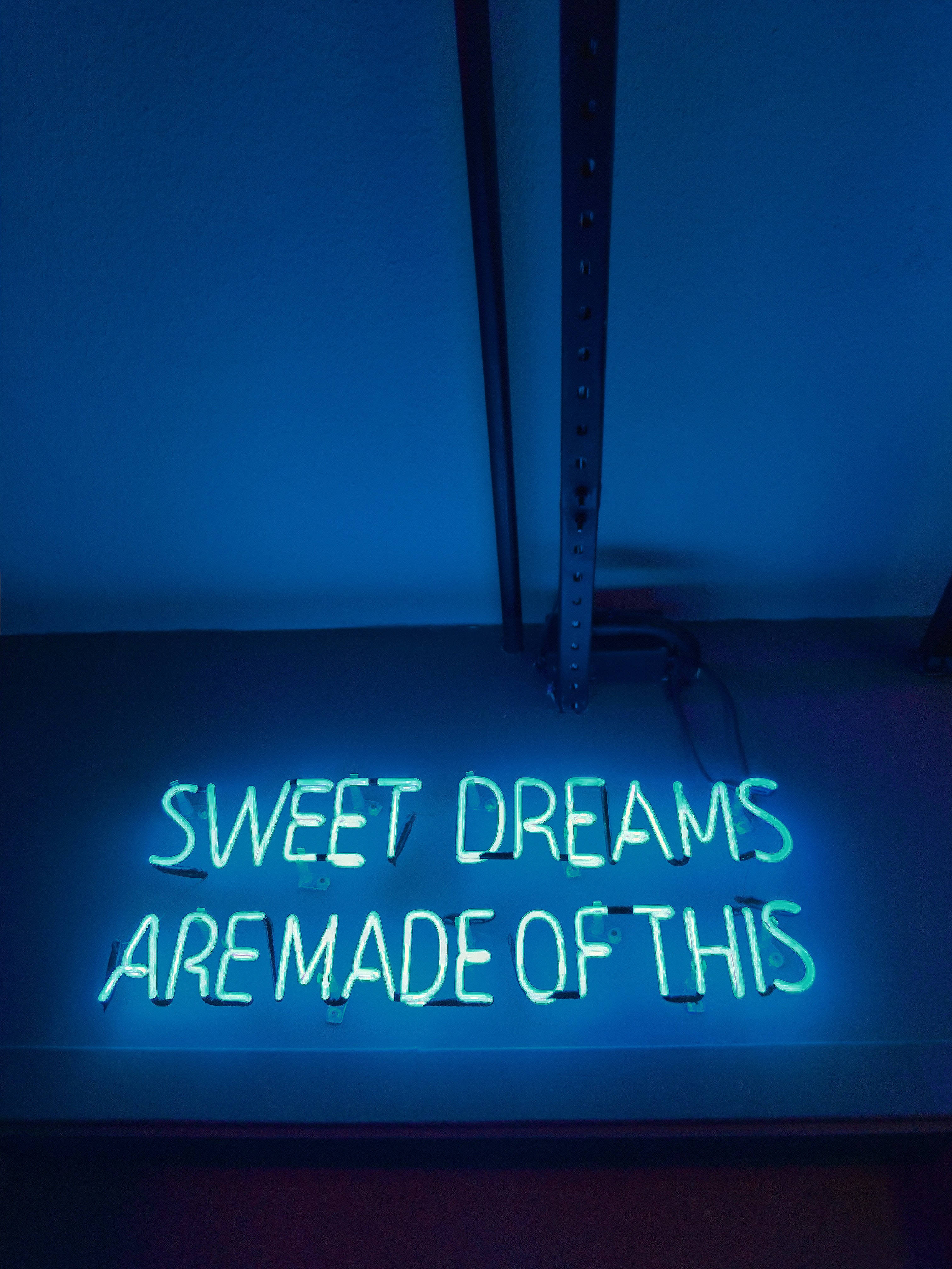 neon sign 桌布 壁紙 — 一個發光的霓虹燈牌寫著 sweet dreams are made of this，在黑暗的牆壁和金屬架上投射出藍光。