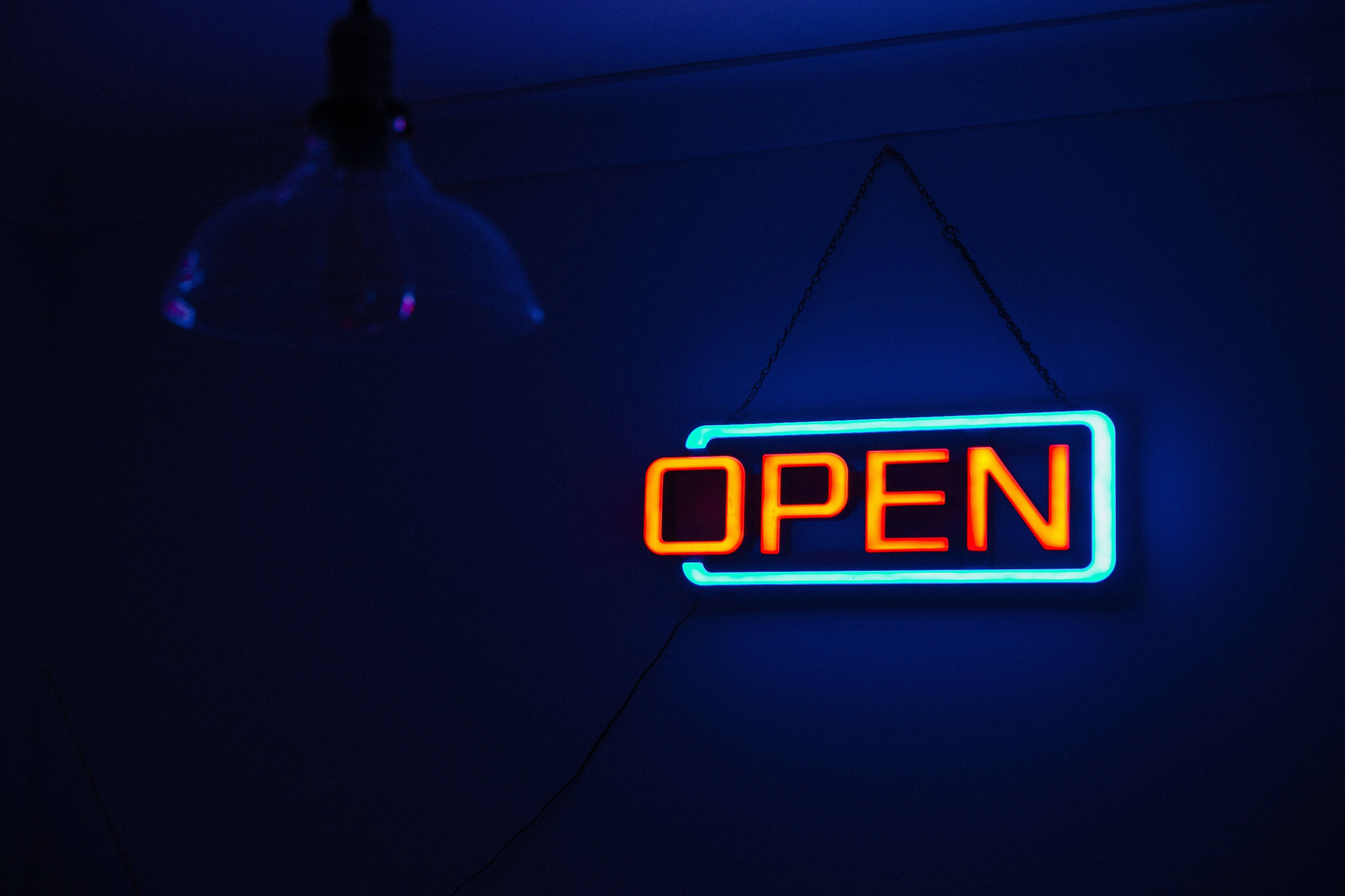 Open 桌布 壁紙 — 一個發光的霓虹燈開放標誌，帶有橙色字母和藍色邊框，懸掛在玻璃吊燈下光線昏暗的房間內。