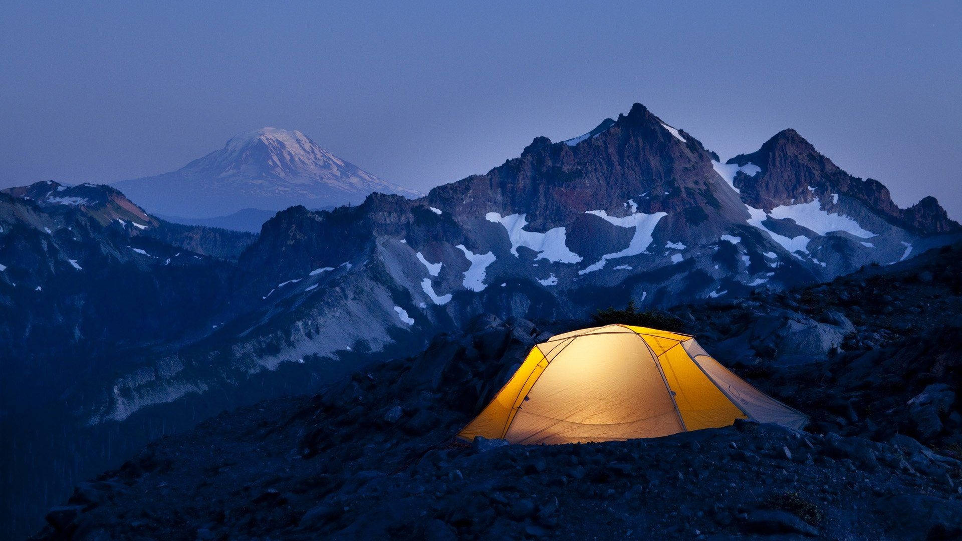Camping 桌布 壁紙 — 一個發光的黃色帳篷，坐落在黃昏時分的岩石山脊上，背後是白雪覆蓋的山峰。