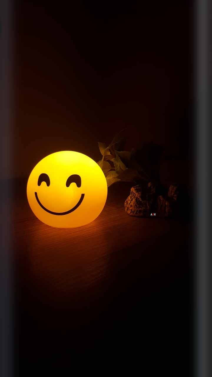 笑臉 桌布 壁紙 — 一個發光的黃色笑臉燈在黑暗的房間中，將溫暖的光線投射在木質表面和旁邊的乾葉子上。