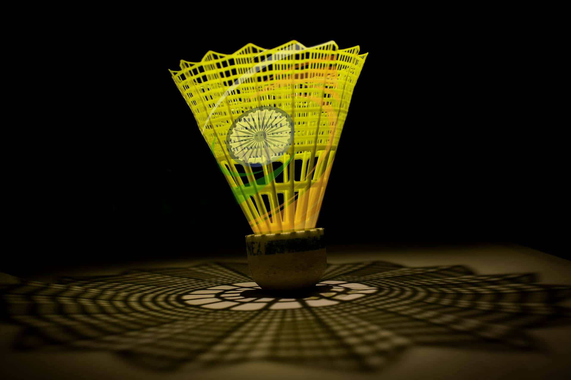 Best Badminton 桌布 壁紙 — 一個發光的黃色羽毛球在戲劇性的燈光下，在黑暗的表面上投射出複雜的幾何陰影。