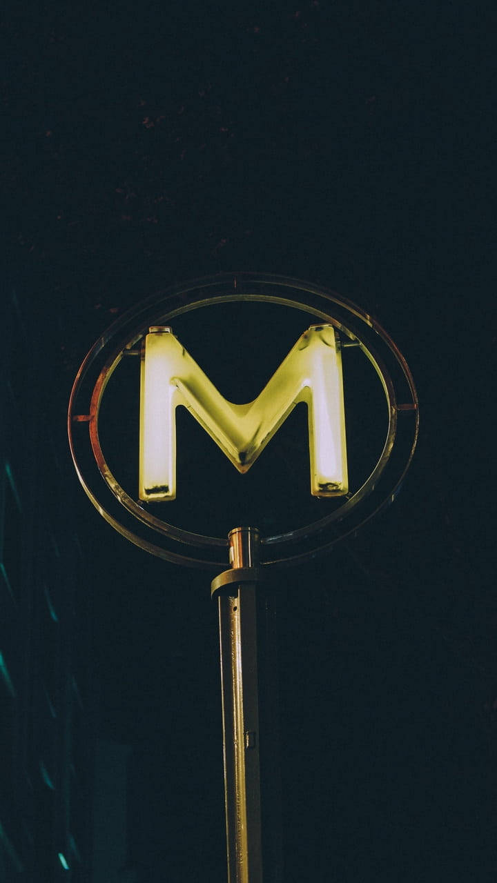 m 桌布 壁紙 — 一個發光的黃色霓虹字母M在圓形框架中，高高聳立在黑暗的夜空下。