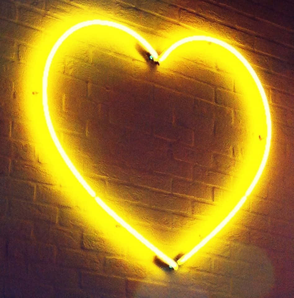 Orange Heart 桌布 壁紙 — 一個發光的黃色霓虹心形在昏暗的空間中，將溫暖的光線投射在紋理磚牆上。
