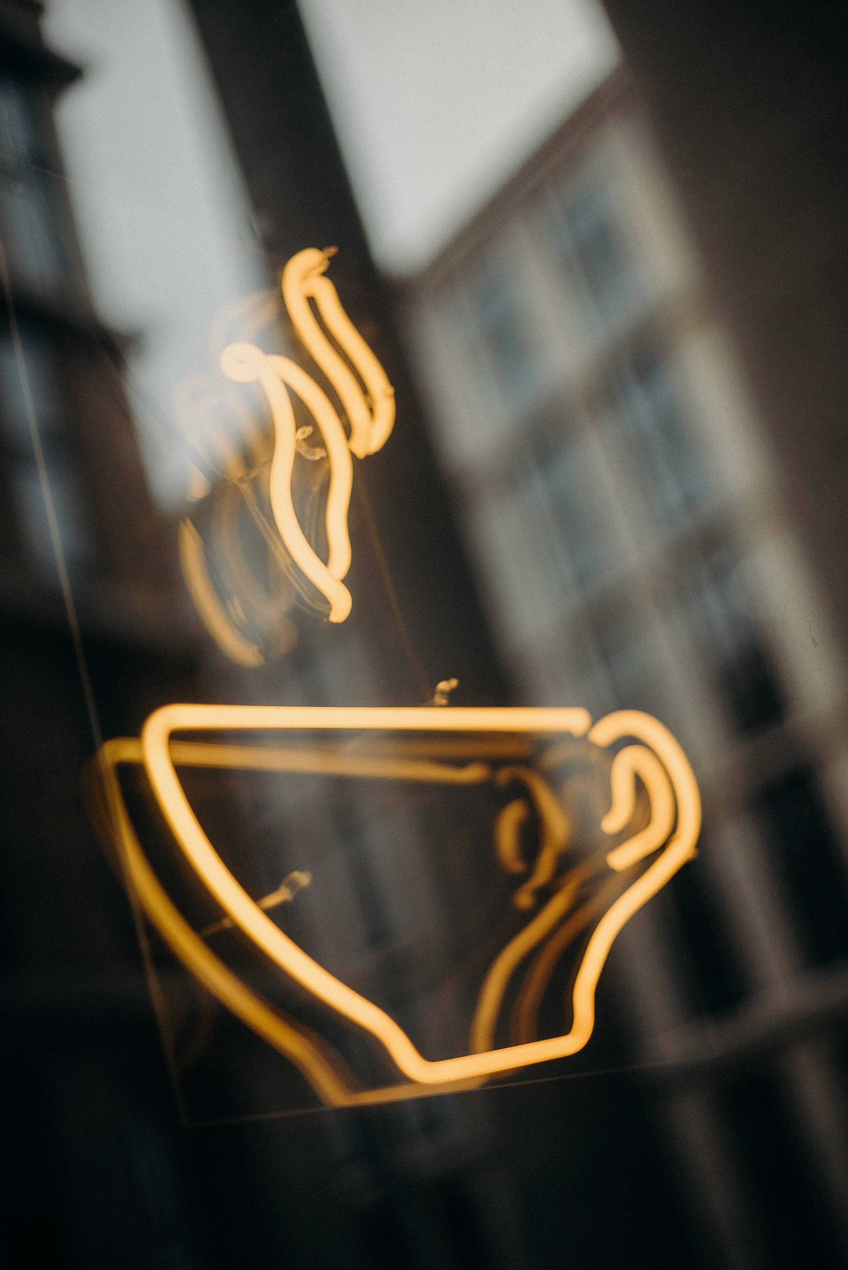 Coffee Shop 桌布 壁紙 — 一個發光的黃色霓虹燈咖啡杯標誌，帶有上升的蒸氣，透過反射模糊城市建築的玻璃窗觀看。