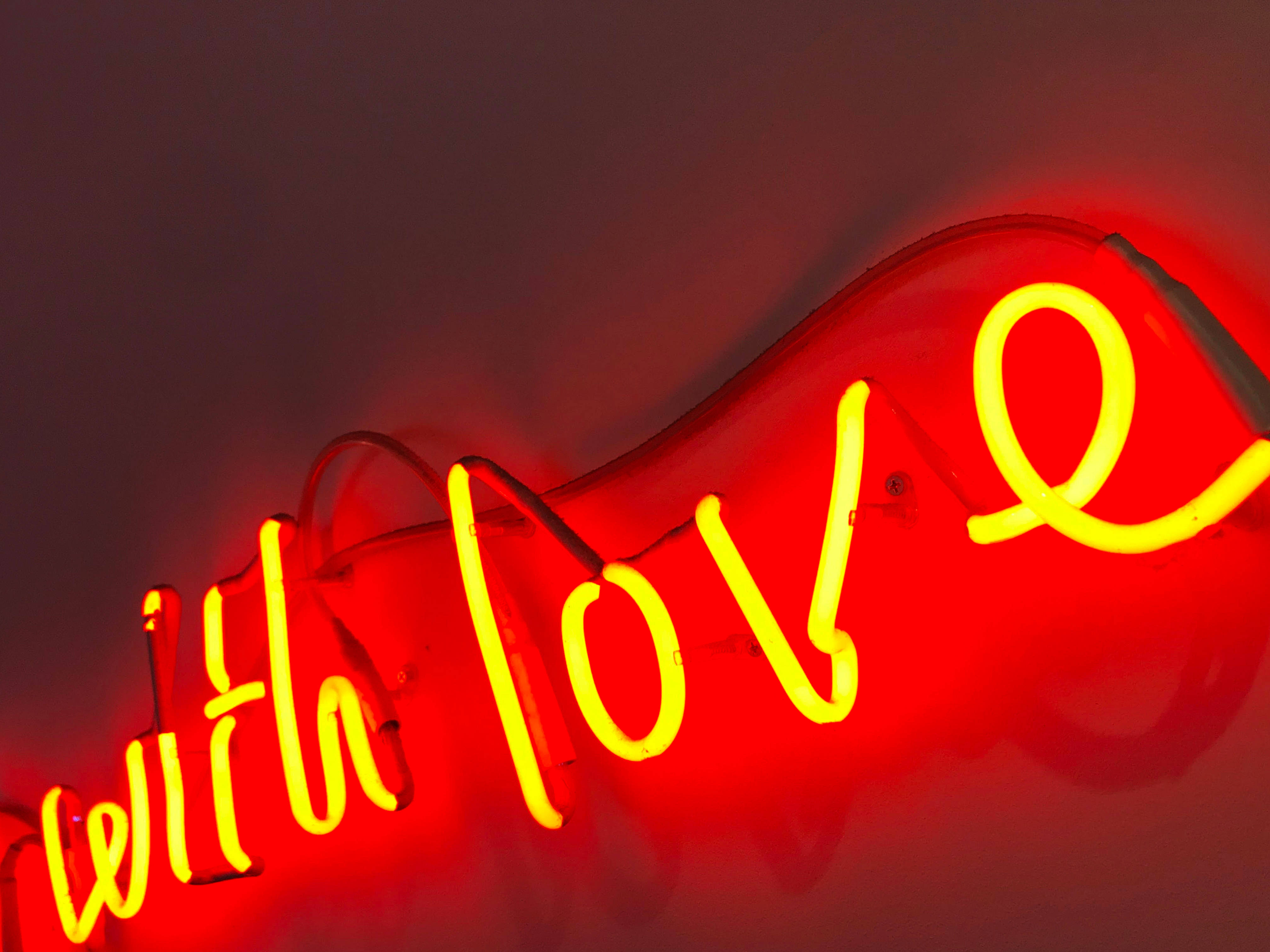 二月 桌布 壁紙 — 一個發光的黃色霓虹燈牌，上面寫著「with love」，在黑暗的牆壁上投射出溫暖的紅色光芒。