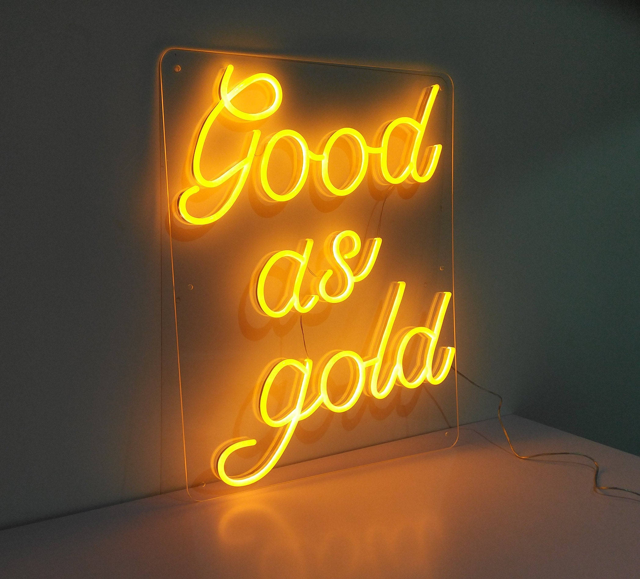 螢光黃 桌布 壁紙 — 一個發光的黃色霓虹燈牌，上面寫著「Good as gold」，在樸素的牆壁和表面上投射出溫暖的光芒。