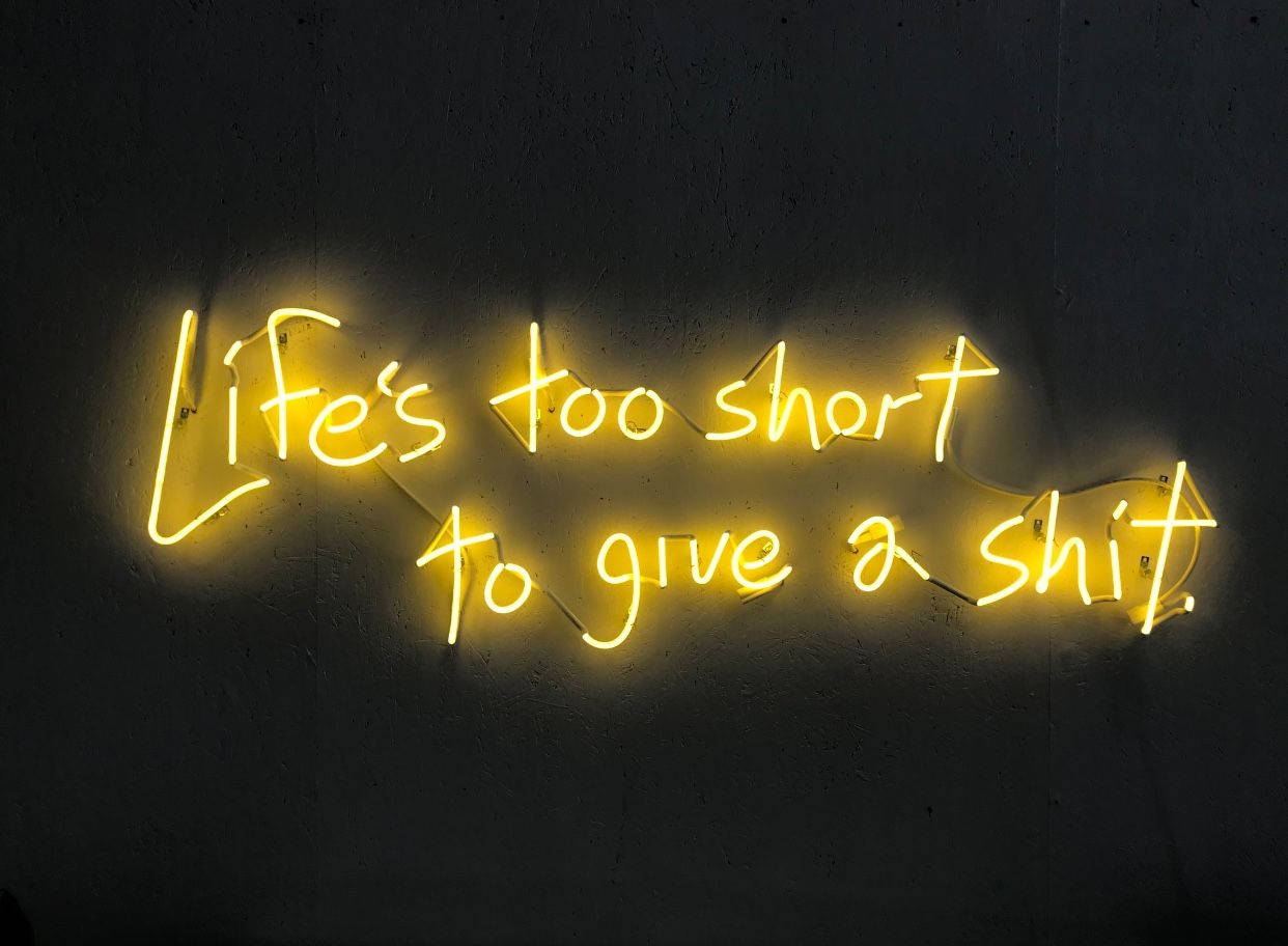 螢光黃 桌布 壁紙 — 一個發光的黃色霓虹燈牌，上面寫著 Life's too short to give a shit，安裝在深色紋理牆上。