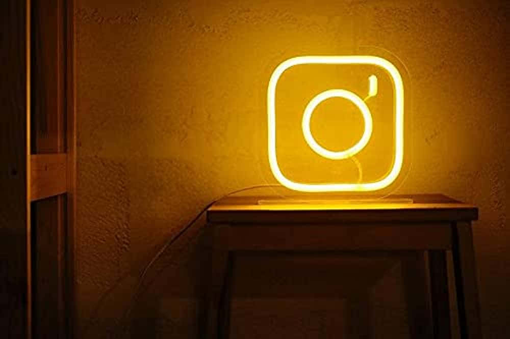Instagram 桌布 壁紙 — 一個發光的黃色霓虹燈牌，形狀像Instagram標誌，放在昏暗房間的木桌上。
