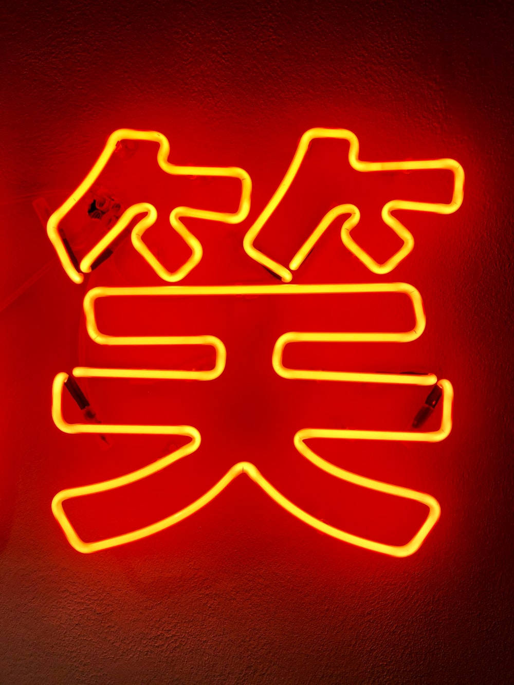  壁紙 — 一個發光的黃色霓虹燈牌，形狀像中文的「笑」字，在紋理牆壁上投射出溫暖的紅光。