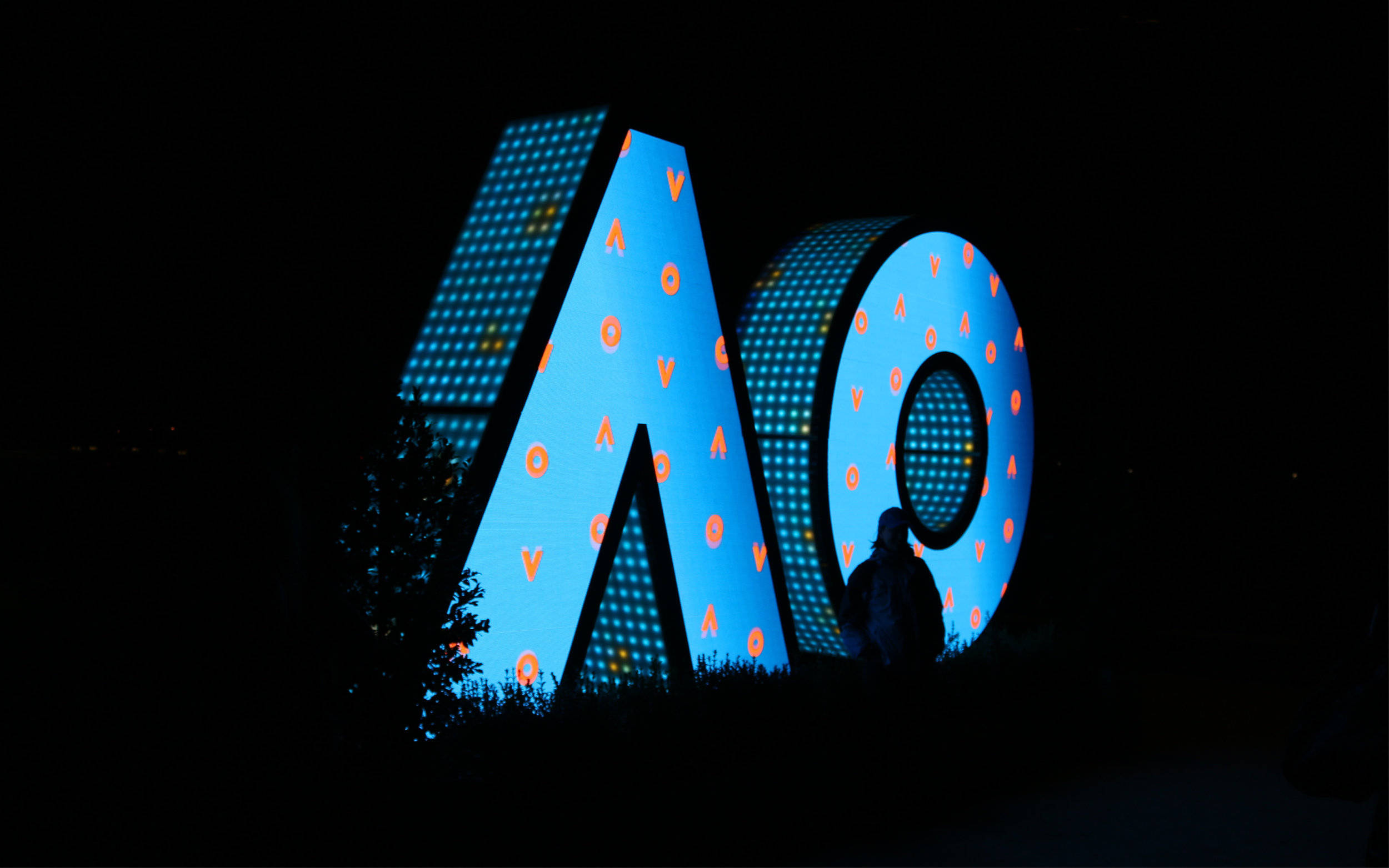 Australian Open 桌布 壁紙 — 一個發光藍色LED字母AO雕塑，帶有橙色圖案，夜晚矗立，旁邊有一個人影。