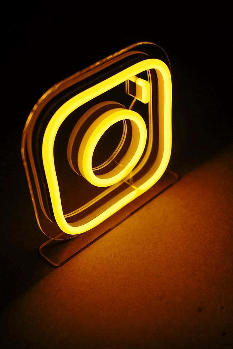 Instagram 桌布 壁紙 — 一個發光黃色霓虹燈牌，形狀像Instagram標誌，在黑暗表面上投射出溫暖的光芒。