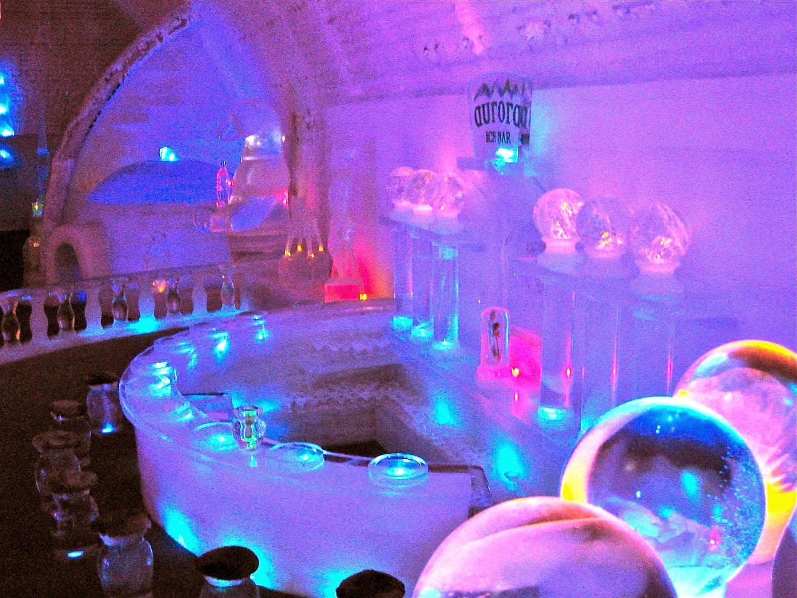 Ice Hotel 桌布 壁紙 — 一個發著藍色和紫色光芒的冰吧，特色是雕刻的冰製家具和環境霓虹燈光下發光的球體。