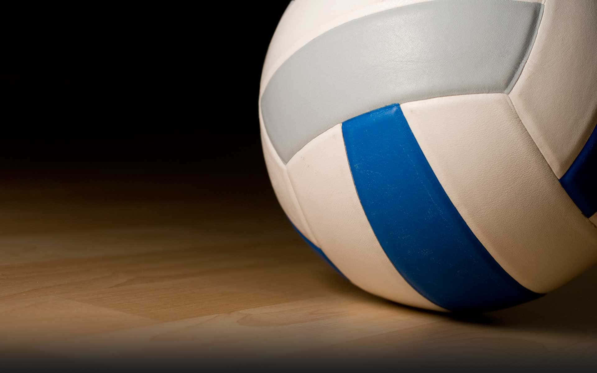 Volleyball Ball 桌布 壁紙 — 一個白色、藍色和灰色的排球特寫，放在木地板上，背景為深色。